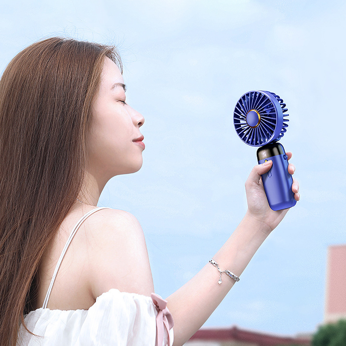 Frau hält einen blauen tragbaren Ventilator. Sie ist draußen und blickt zum Ventilator. Blauer Himmel im Hintergrund.