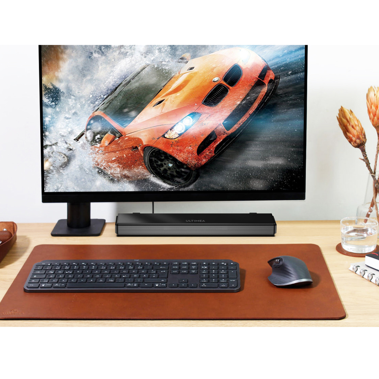 Configuración de escritorio con monitor mostrando un coche, teclado, ratón y alfombrilla de cuero marrón.