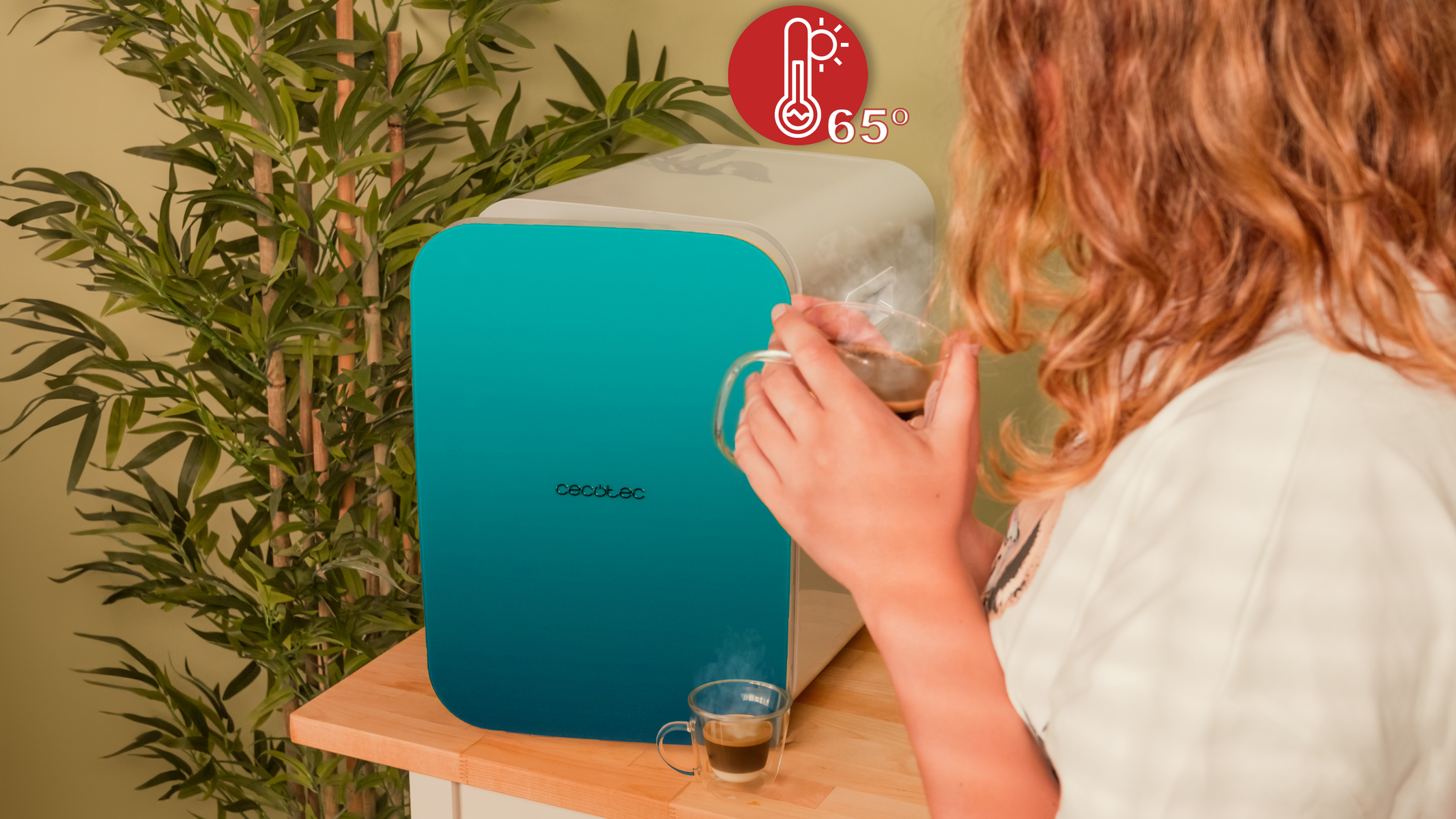 Una mujer con un vaso de café cerca de una cafetera blanca. Un termómetro muestra 65 grados.