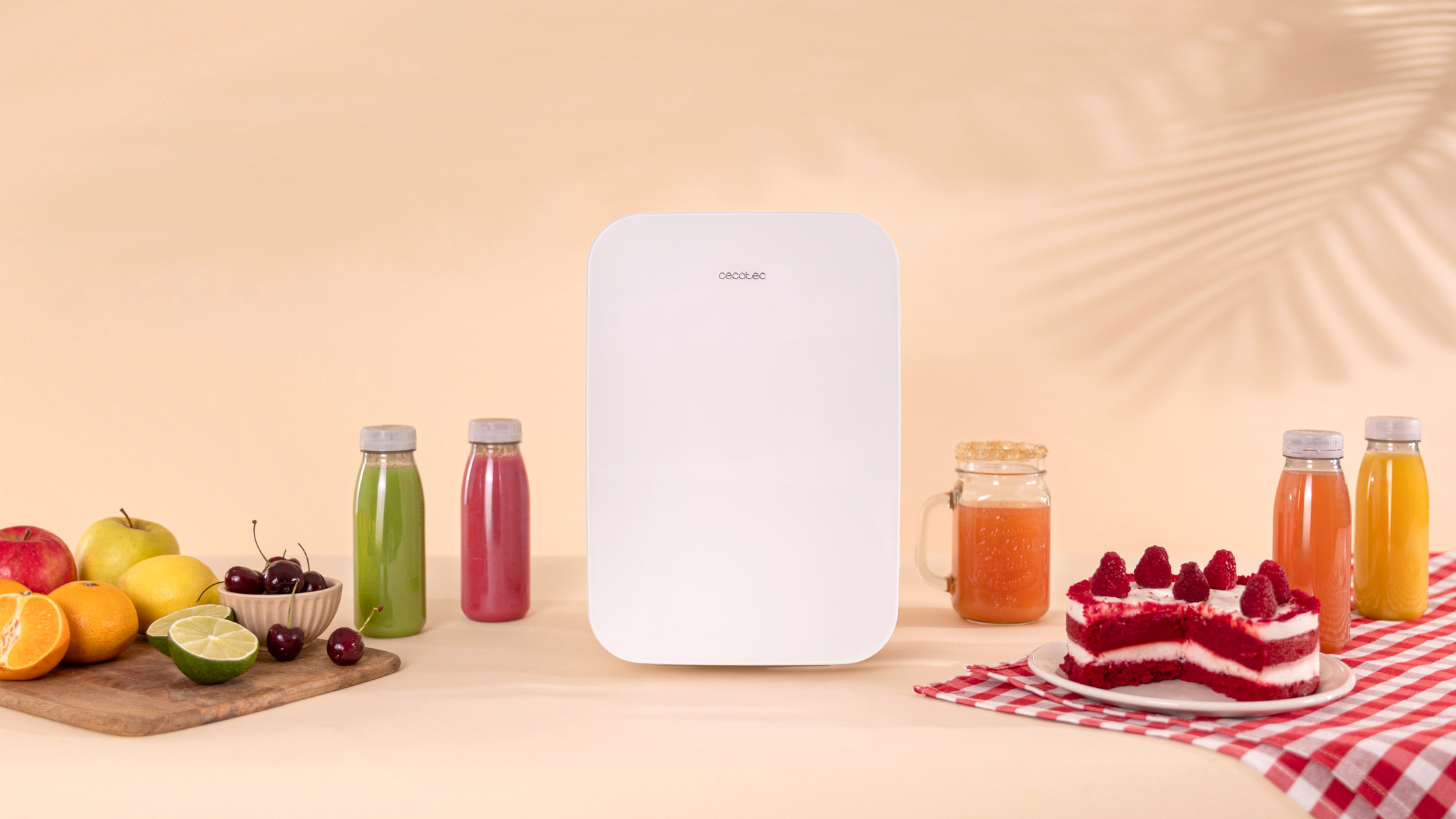 Un mini refrigerador blanco con botellas de jugo, frutas y un pastel. Todo sobre un fondo color melocotón.