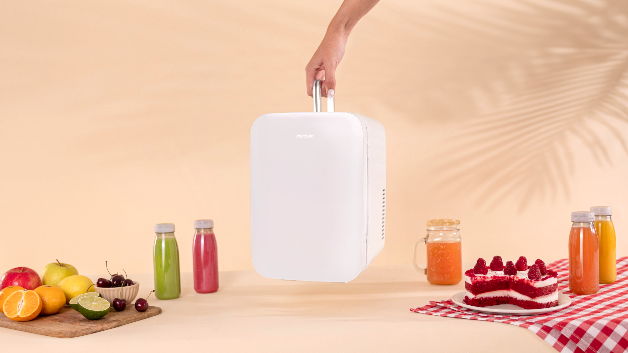 Una mano sosteniendo una mini nevera blanca. Frutas, pastel y botellas de jugo en la mesa.