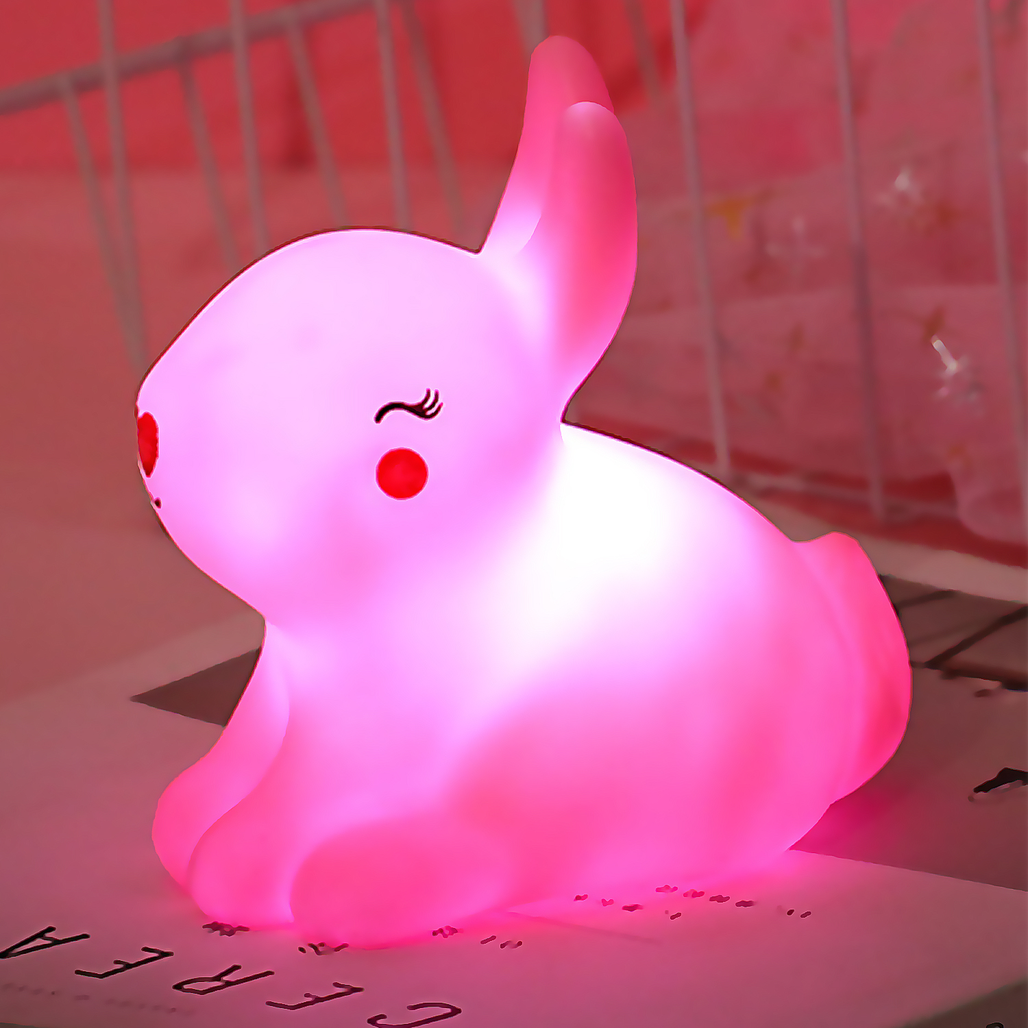Luce notturna a forma di coniglietto rosa con occhi chiusi, seduto su un tavolo. Luce soffusa e brillante.