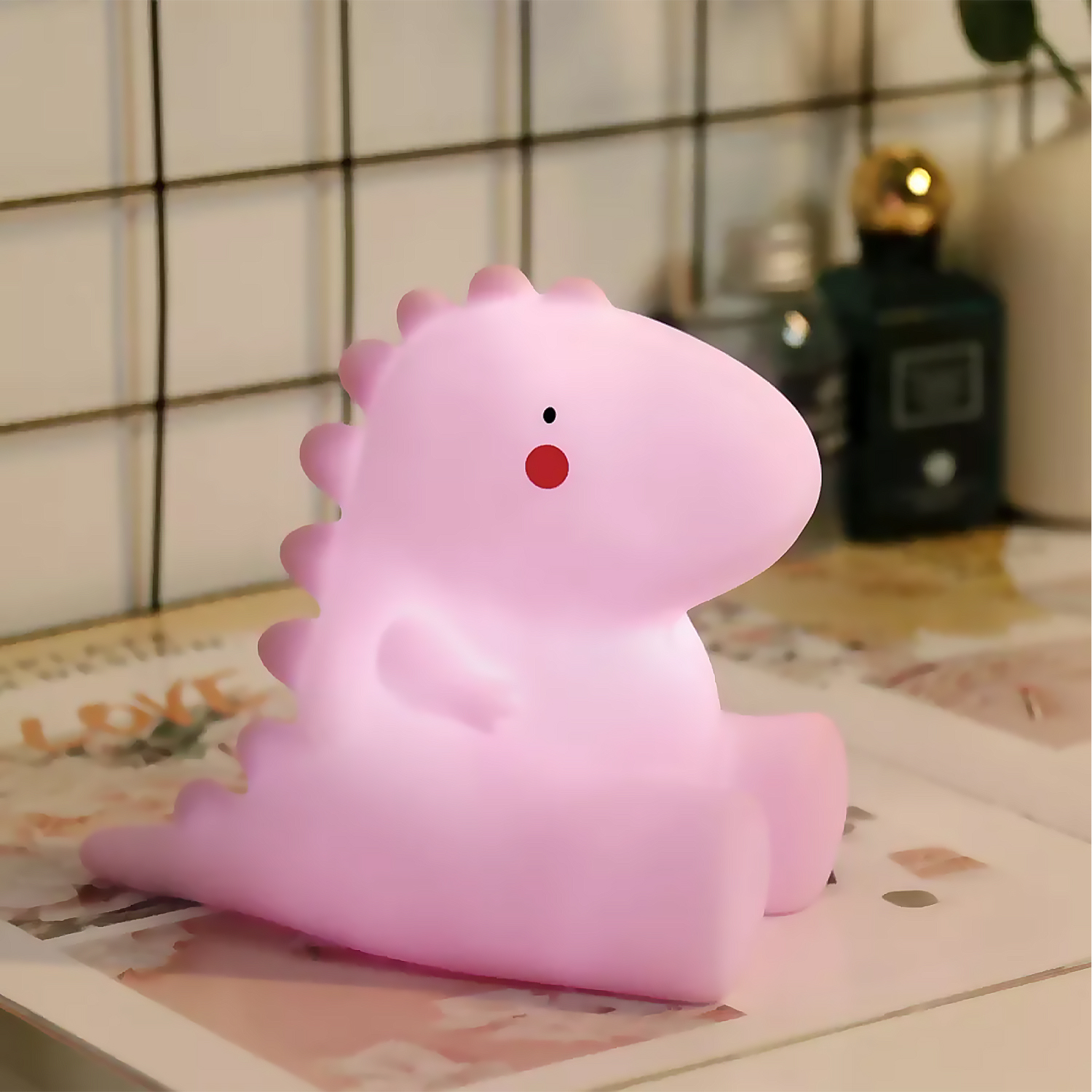 Una luz de noche con forma de dinosaurio rosa. Tiene un punto rojo en la cara y está iluminada.