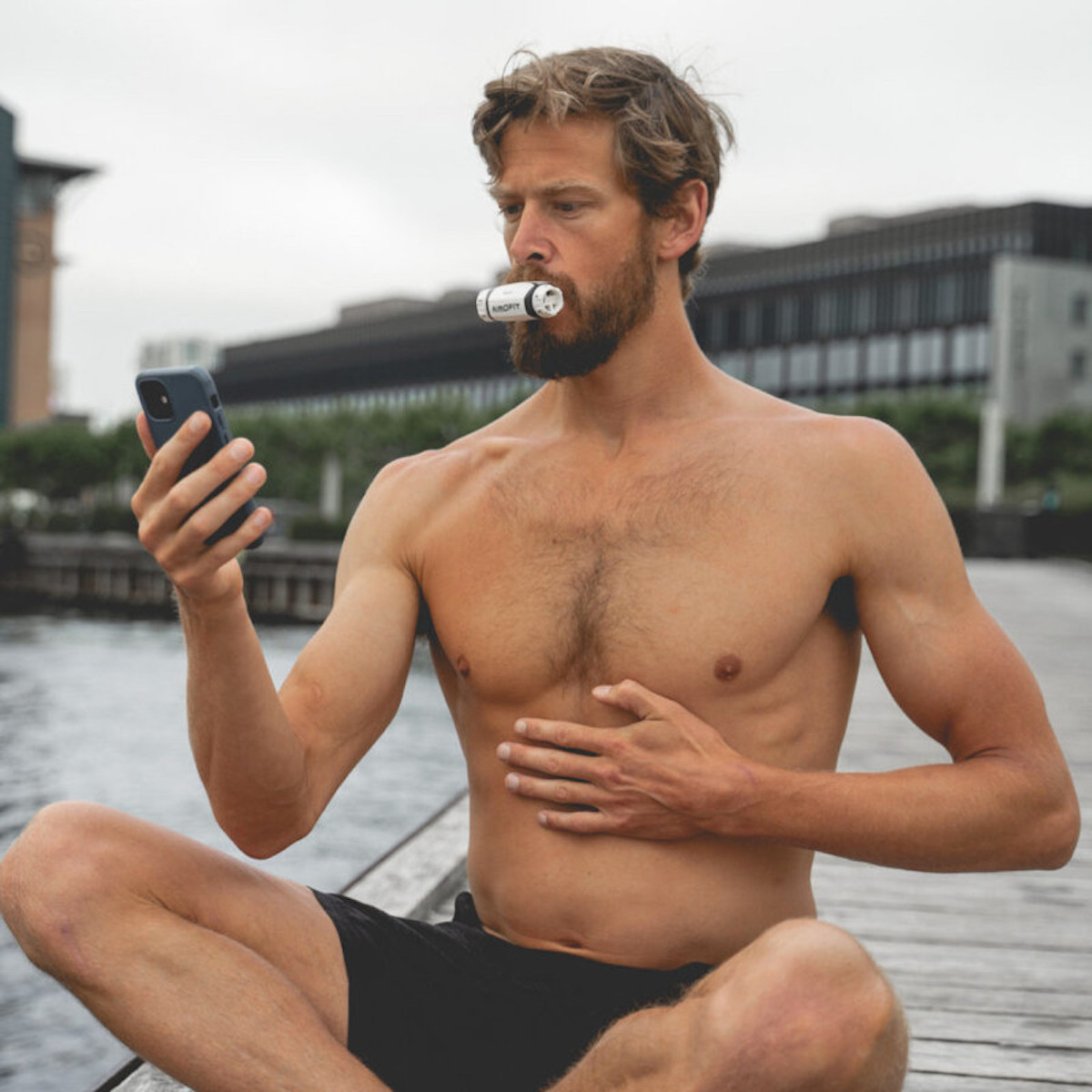 Ein Mann sitzt im Freien und benutzt ein Atemalkoholmessgerät und hält ein Telefon, mit Wasser im Hintergrund.