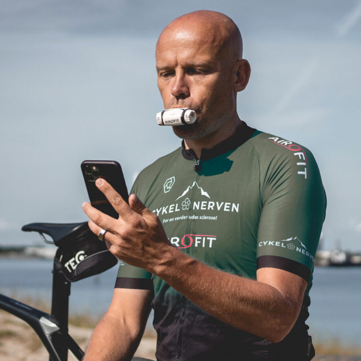 Mann im Sporttrikot, der ein Telefon benutzt, mit einem Gerät im Mund. Küstenhintergrund.