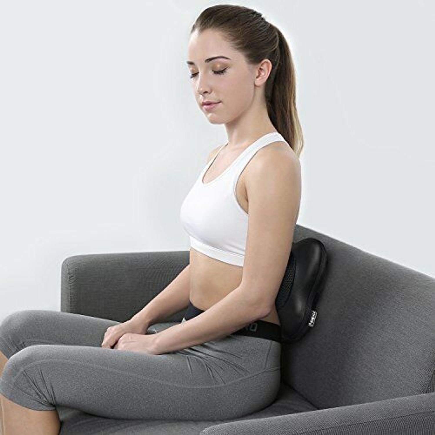 Eine Frau sitzt auf einem grauen Sofa mit einem schwarzen Massagekissen im Rücken.