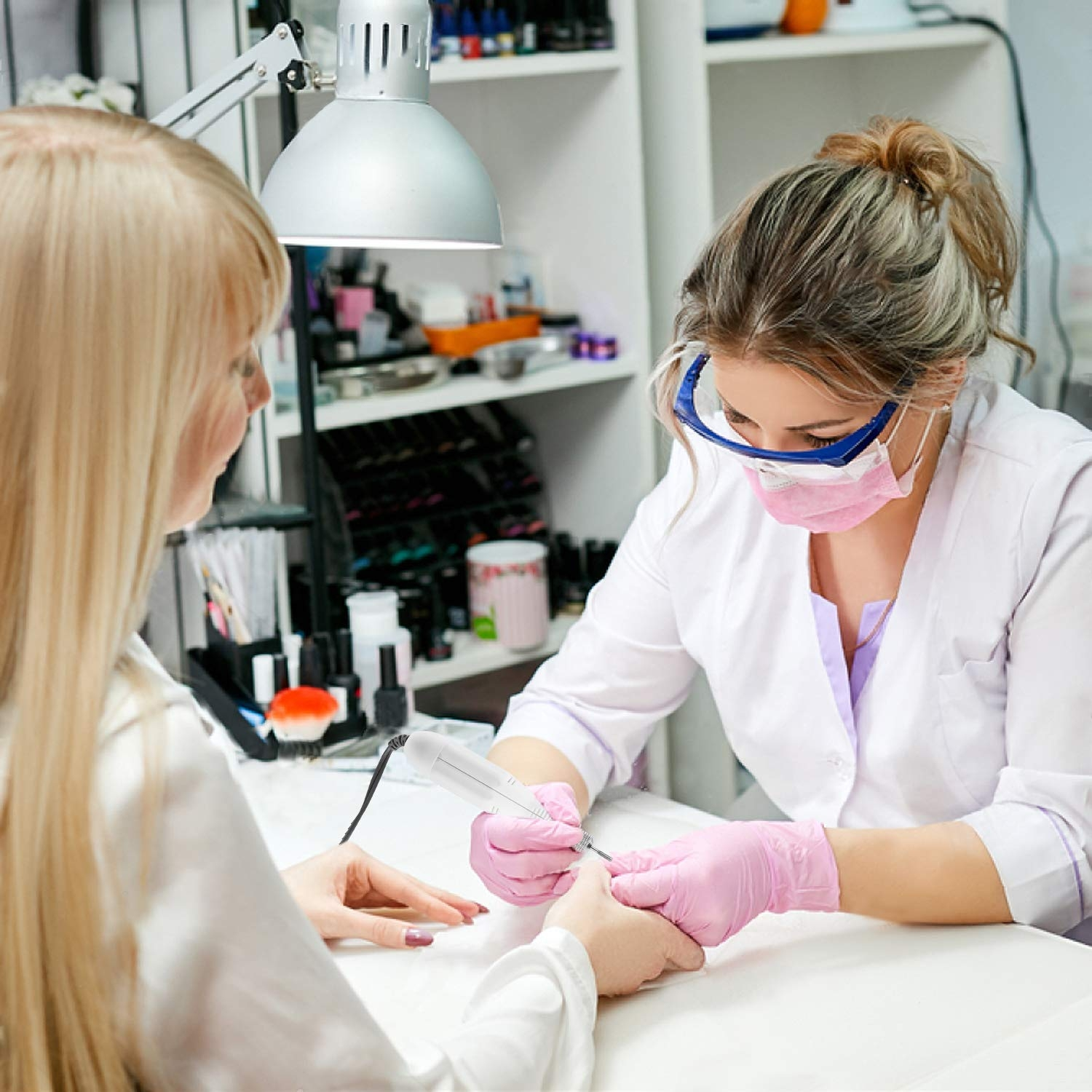 Nageltechnikerin arbeitet an den Nägeln einer Kundin. Sie trägt Handschuhe und Maske, Lampe und Tisch.