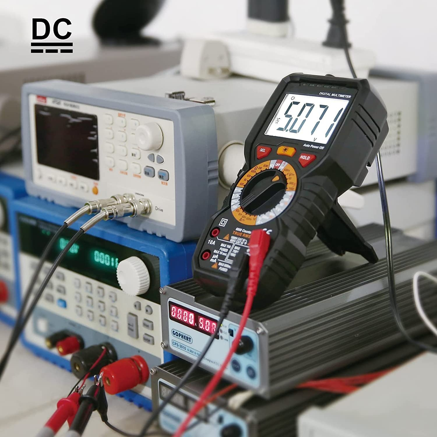 Digitalmultimeter mit schwarzem Gehäuse, Anzeige zeigt 5,071, mit roten und schwarzen Sonden, und andere Geräte im Hintergrund.