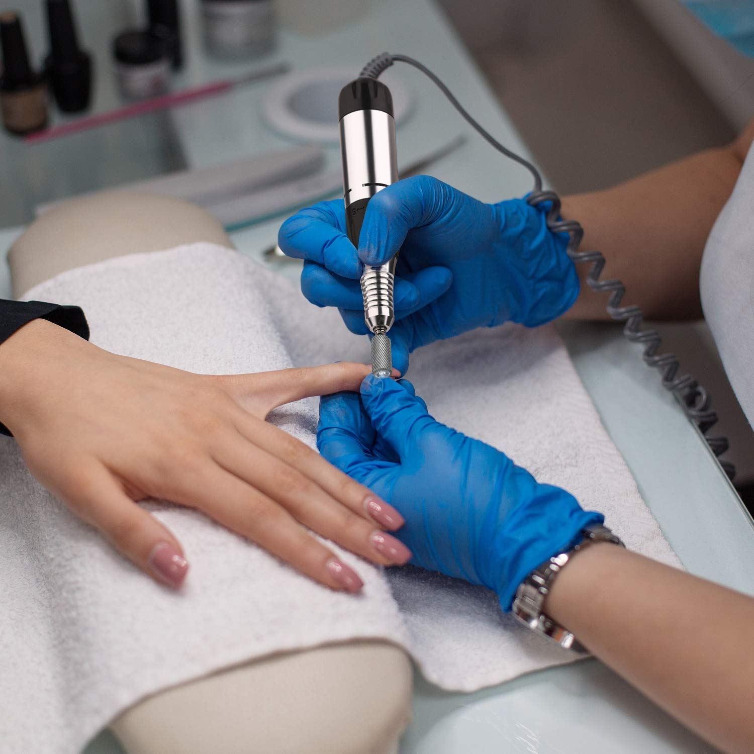 Eine Maniküristin, die einen Nagelbohrer an einem Fingernagel des Kunden verwendet. Der Kunde trägt blaue Handschuhe.