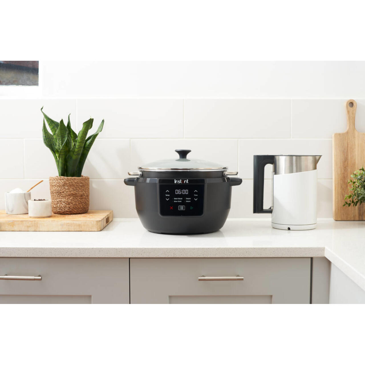 Zwarte Instant Pot op een aanrecht, met een plant en andere keukenartikelen. Witte tegelachtergrond.