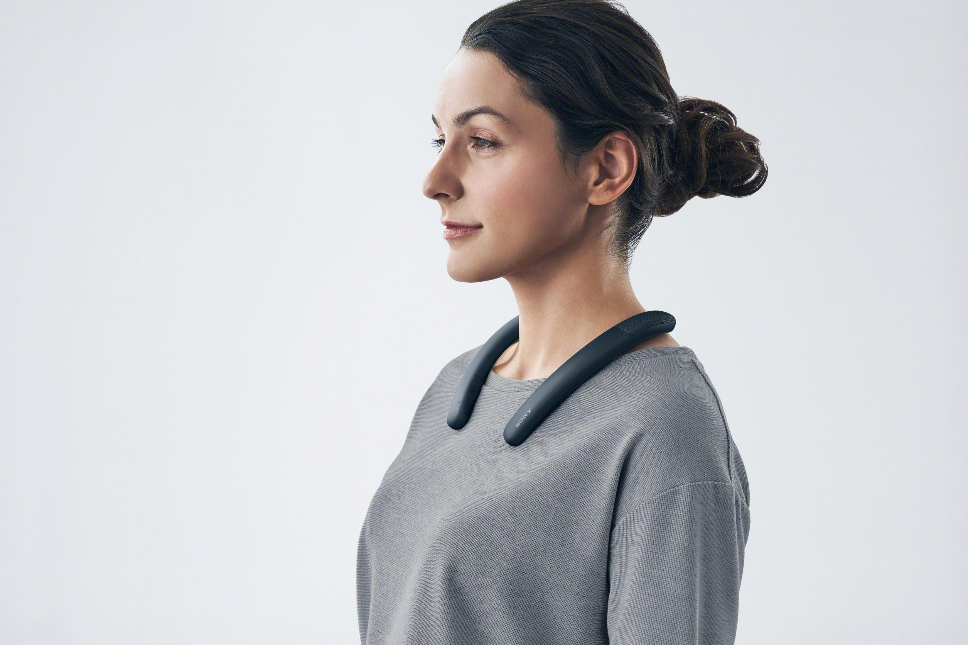 Kobieta w czarnych słuchawkach na szyję, szarej koszuli i na jasnym tle. Ujęcie z profilu.