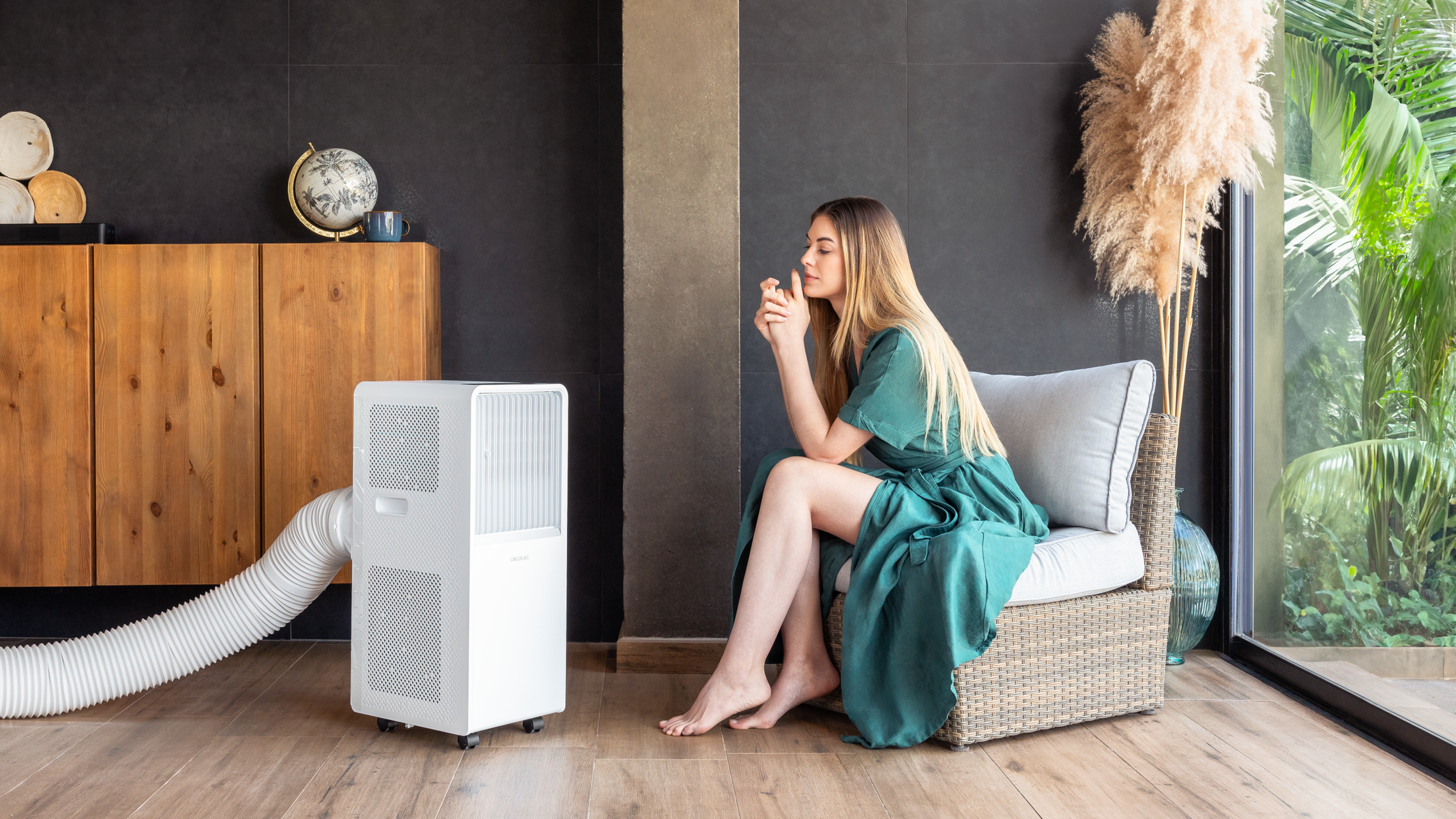 Kobieta w zielonej sukience siedzi obok białego przenośnego klimatyzatora. Tło z drewnianą szafką i roślinami.