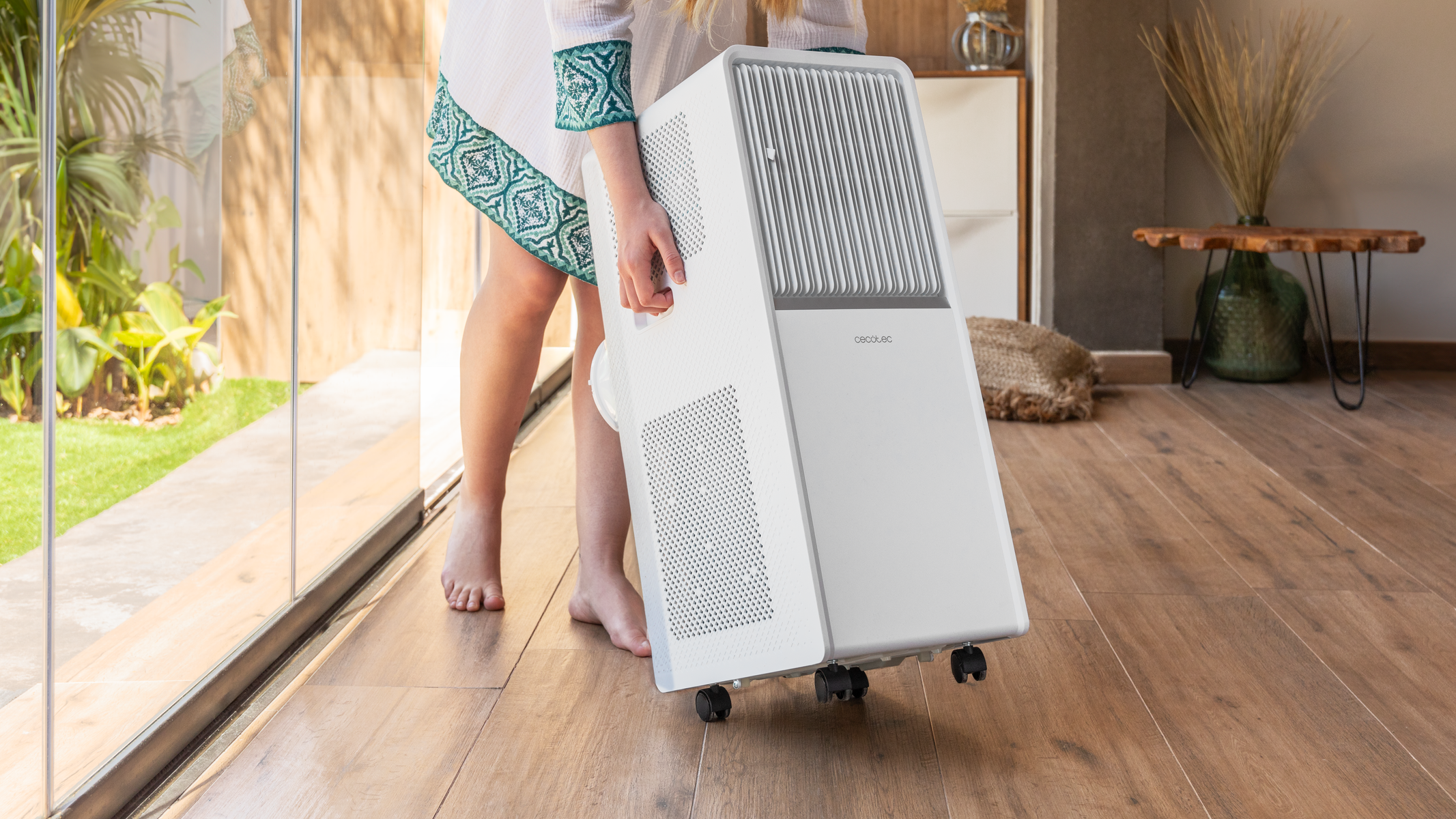Osoba trzymająca białą przenośną klimatyzację, idąca w pomieszczeniu po drewnianej podłodze.