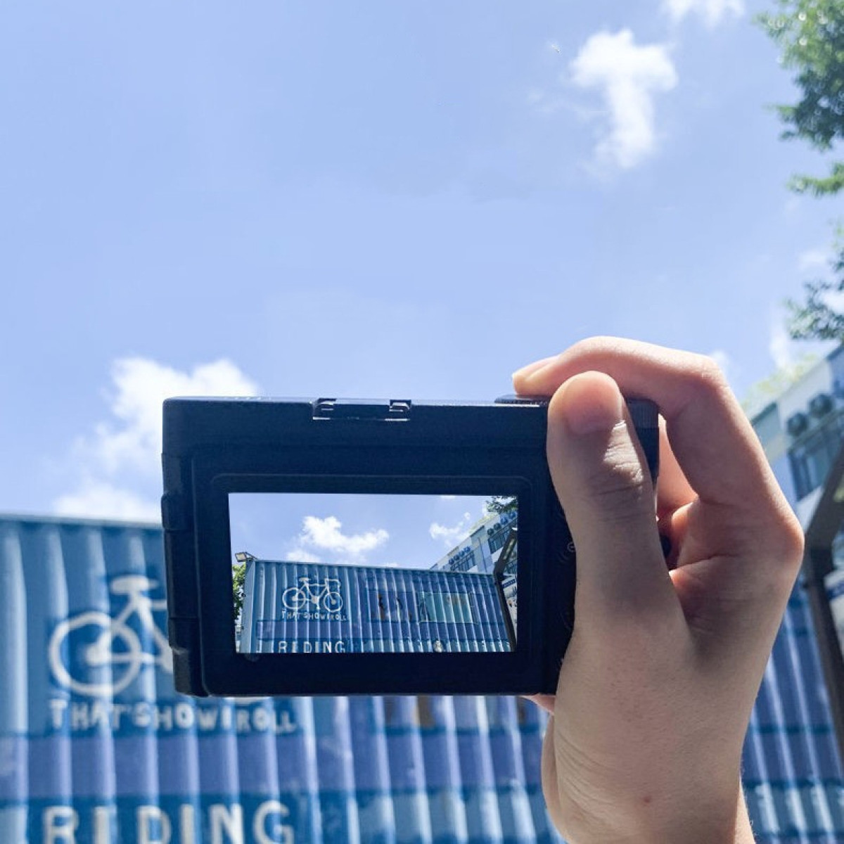 Dłoń trzyma aparat, ekran pokazuje budynek. Niebieskie tło kontenera, niebieskie niebo z chmurami.