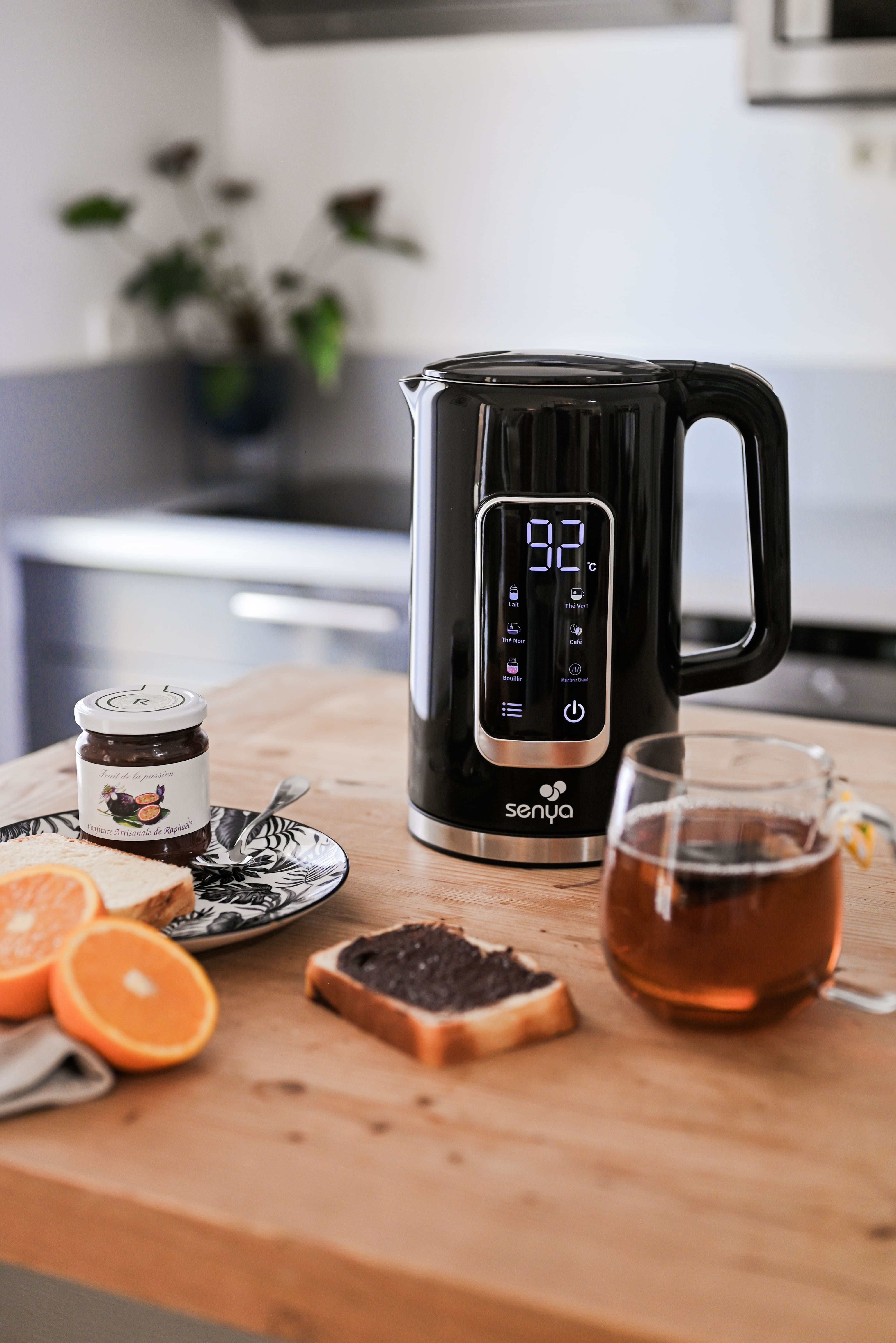 Frühstücksszene: Schwarzer Wasserkocher, Toast mit Aufstrich, Orangenscheiben, Tee im Glas und Marmeladenglas auf Holztisch.