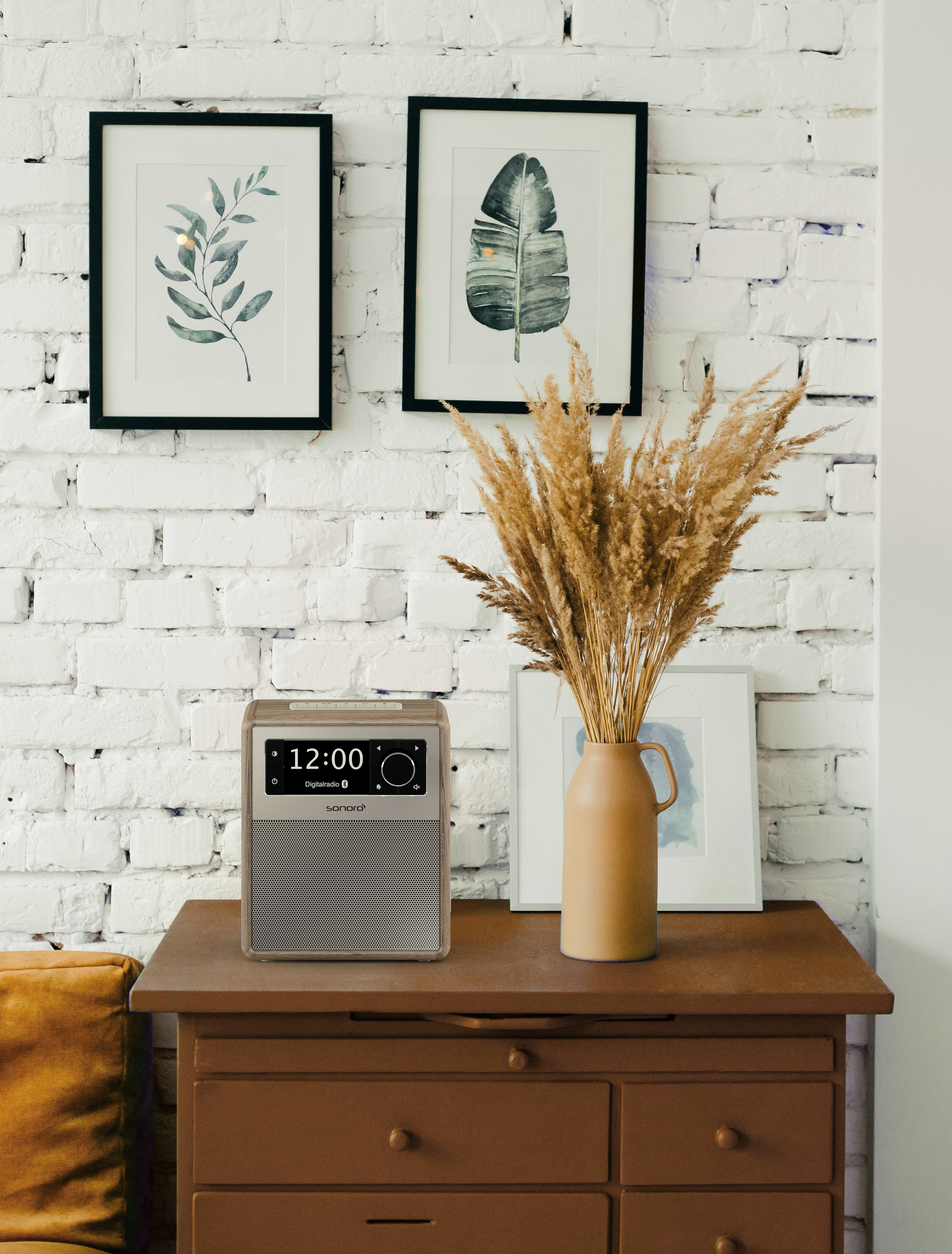 Braune Holzkommode mit Radio. Zwei gerahmte Bilder und eine Vase mit getrockneten Blumen.