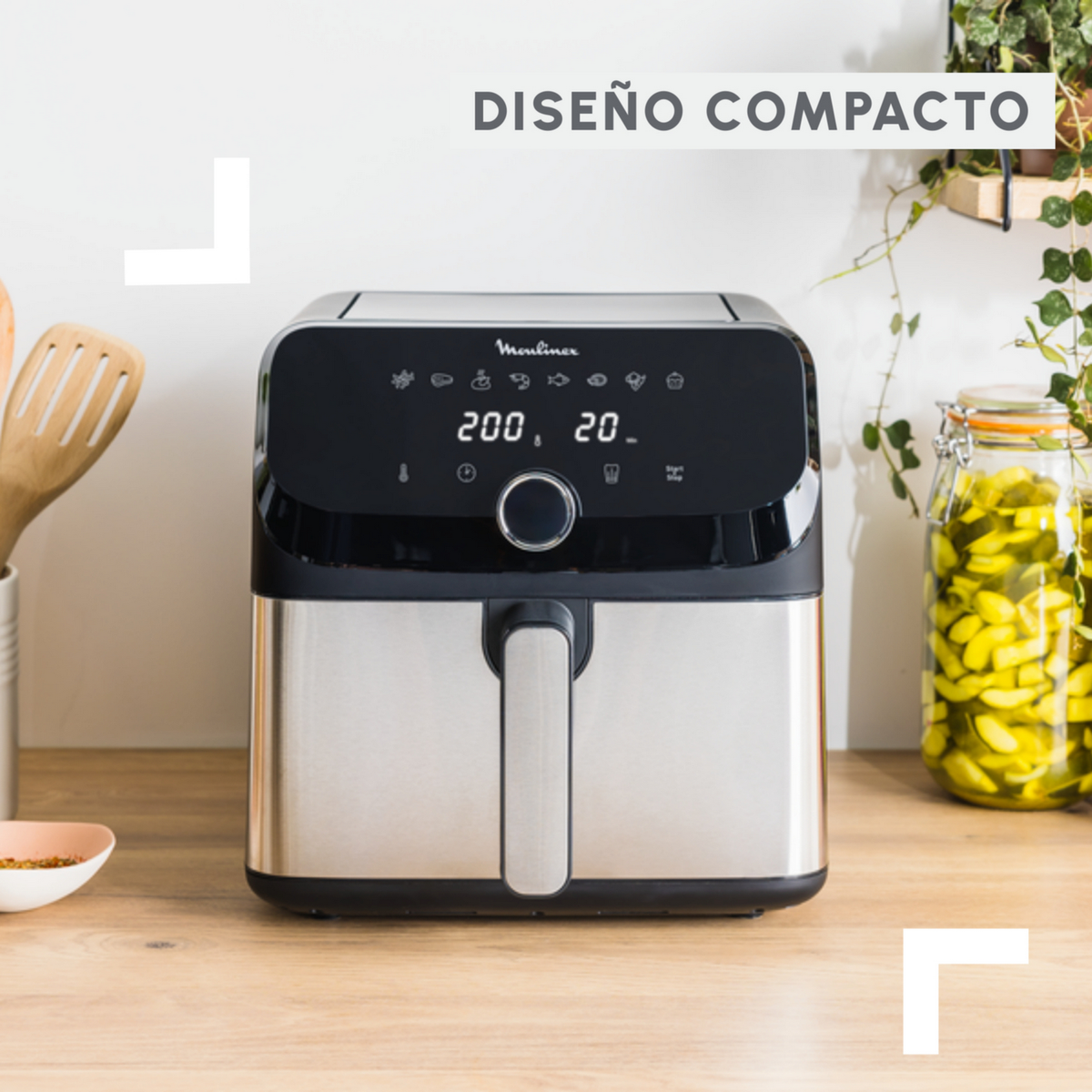 Frytkownica beztłuszczowa Moulinex na drewnianym stole. Srebrna i czarna z wyświetlaczem, słoikami i przyborami kuchennymi.