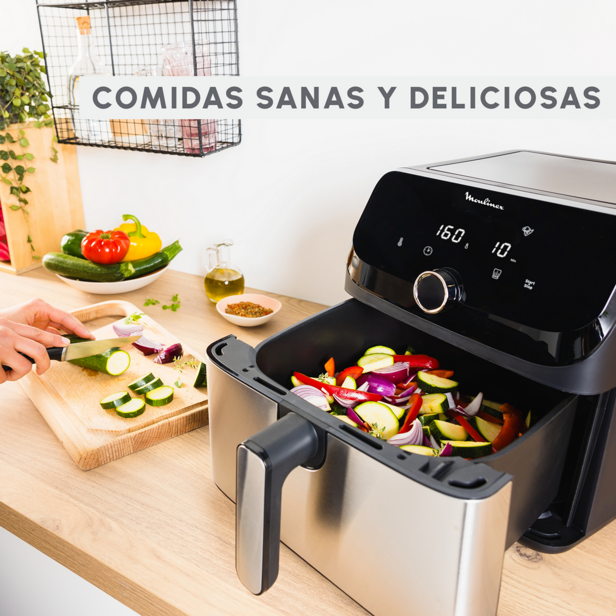 Frytkownica powietrzna z warzywami w środku. Ręka kroi cukinię. Drewniany stół. Białe tło.