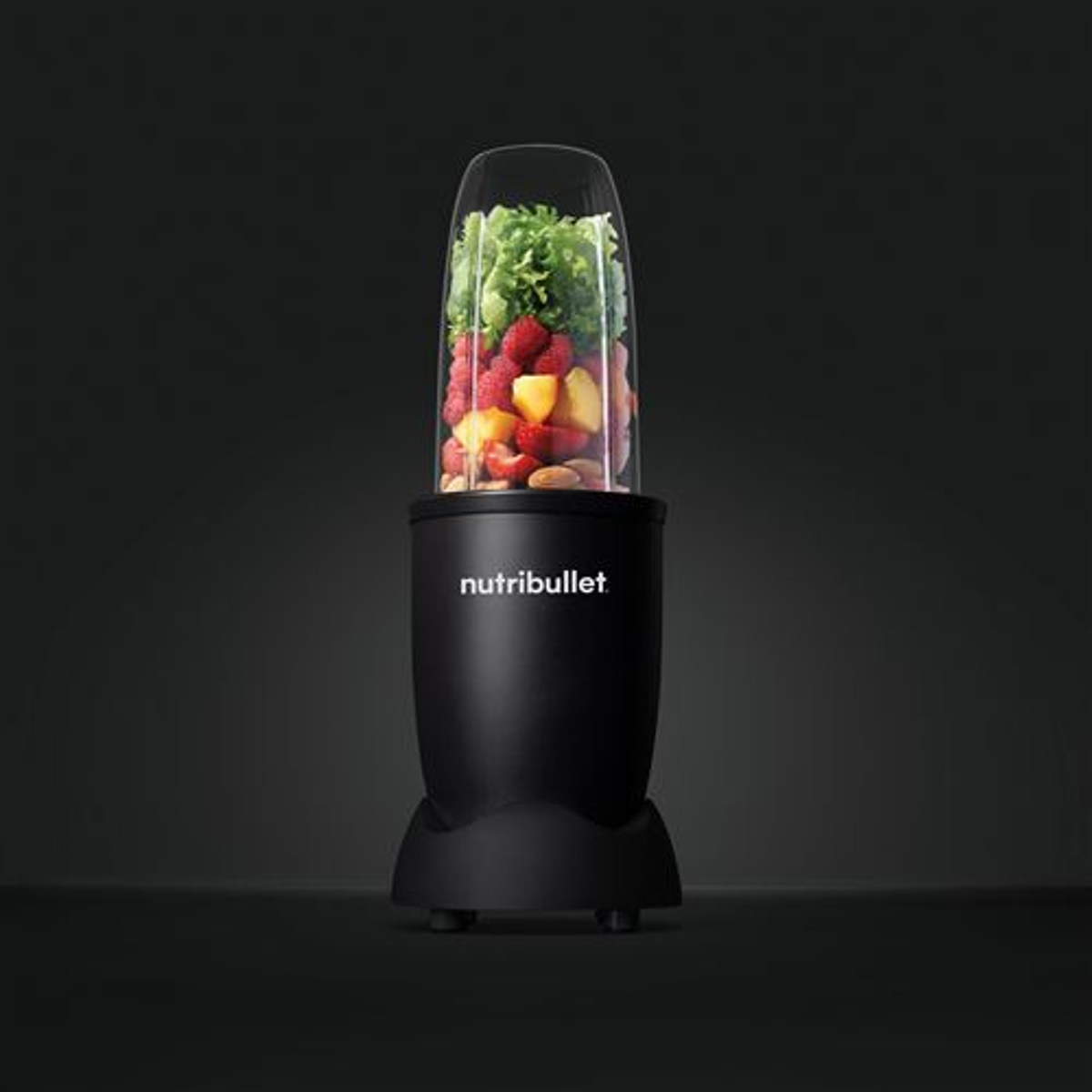 Una licuadora NutriBullet negra llena de frutas y verduras. Fondo negro.