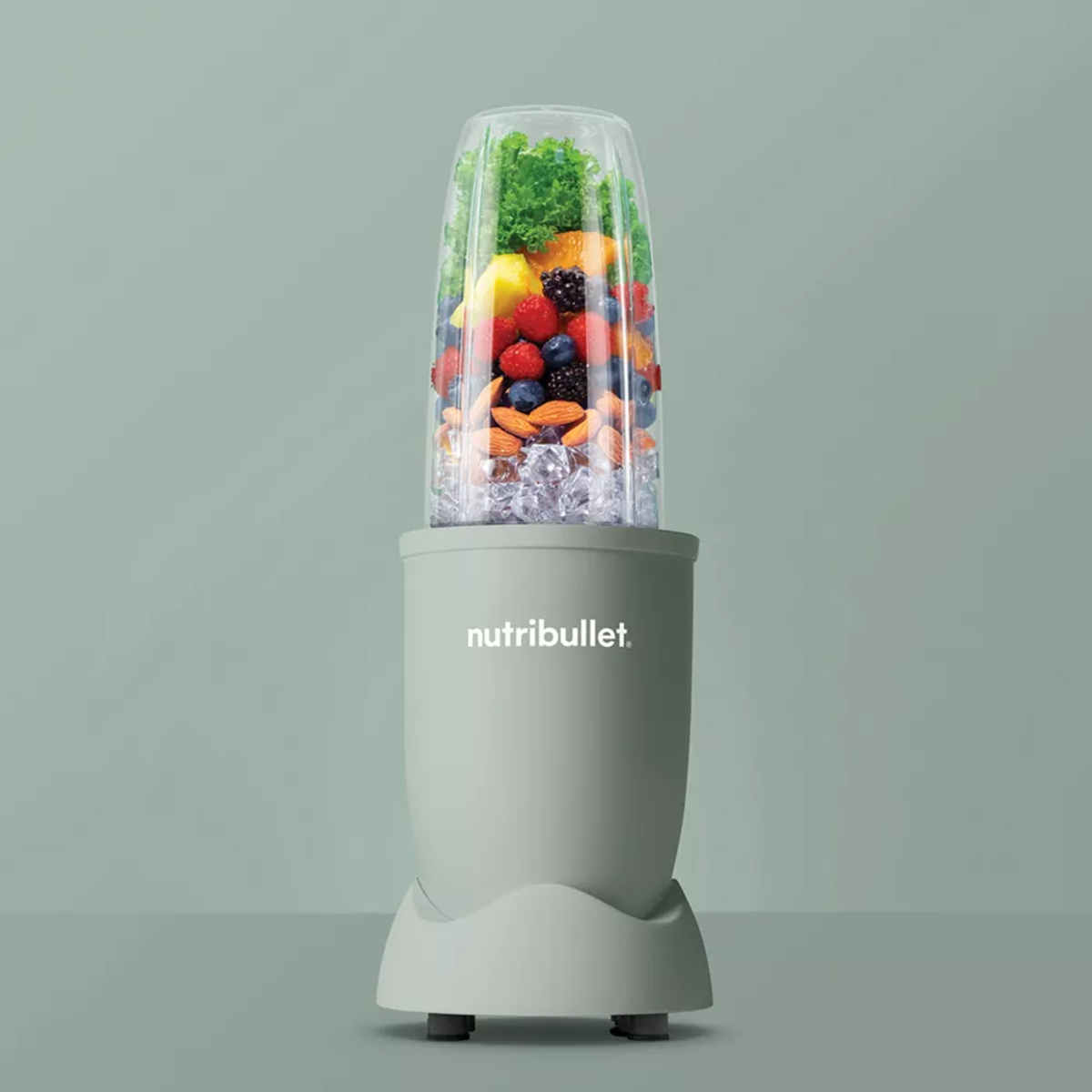 Una licuadora con un recipiente transparente lleno de frutas, hielo y verduras, sobre un fondo verde.