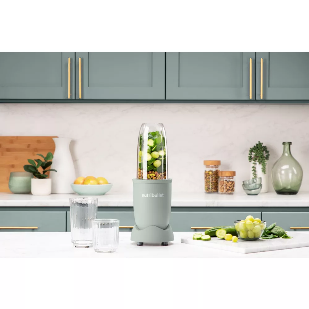 Una licuadora NutriBullet verde con ingredientes dentro, vasos y una tabla de cortar en una superficie blanca.