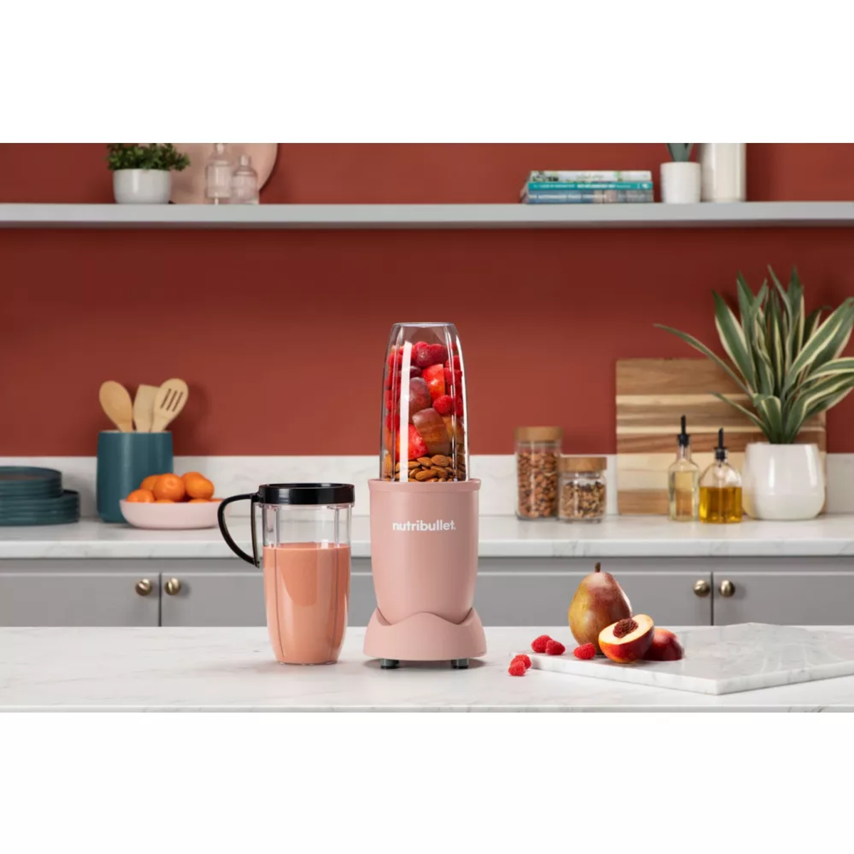 Różowy blender Nutribullet z owocami w środku, kubek z napojem i owoce na blacie.