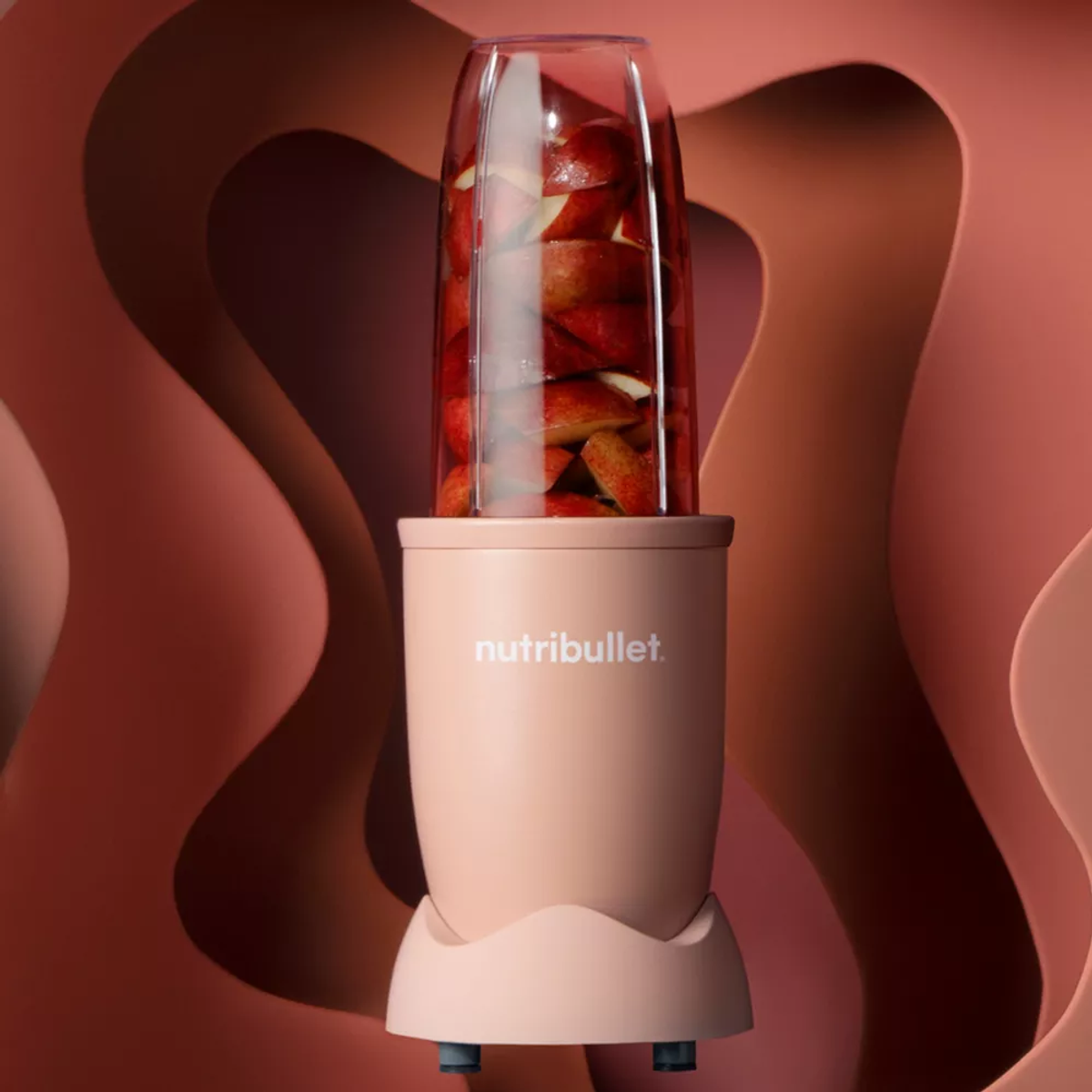 Różowy blender Nutribullet z plasterkami jabłek w środku. Tło ma różowy, warstwowy wzór.