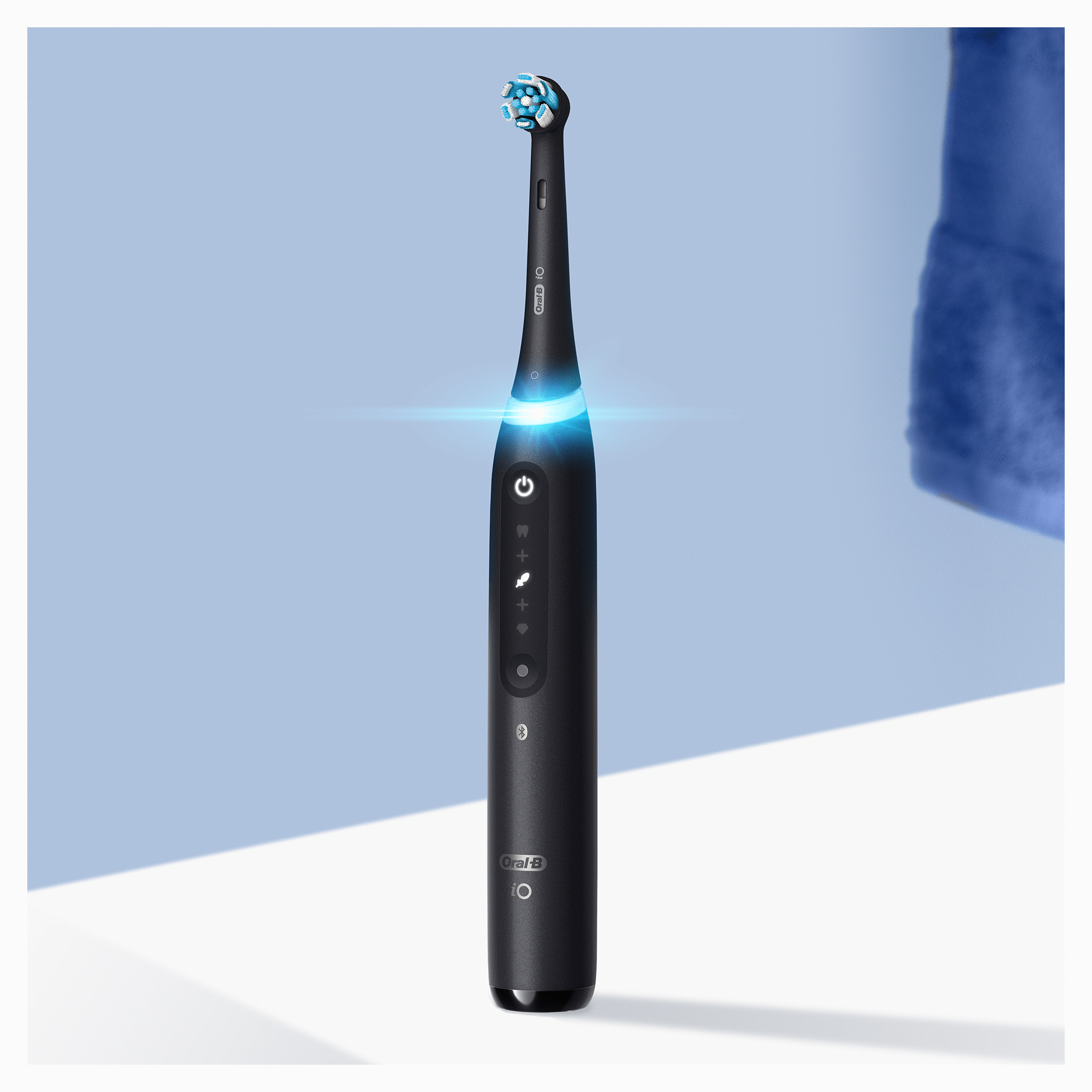 Schwarze elektrische Zahnbürste mit blauem Licht, auf weißer Oberfläche mit hellblauem Hintergrund, blaues Handtuch rechts.