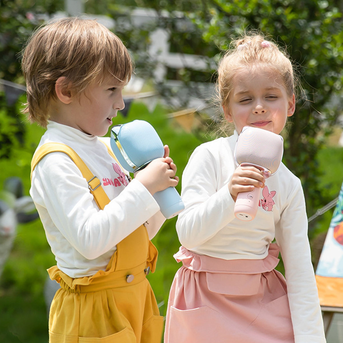Zwei Kinder halten Mikrofone. Eines in gelben Overalls, das andere in rosa Rock. Grüner Hintergrund.