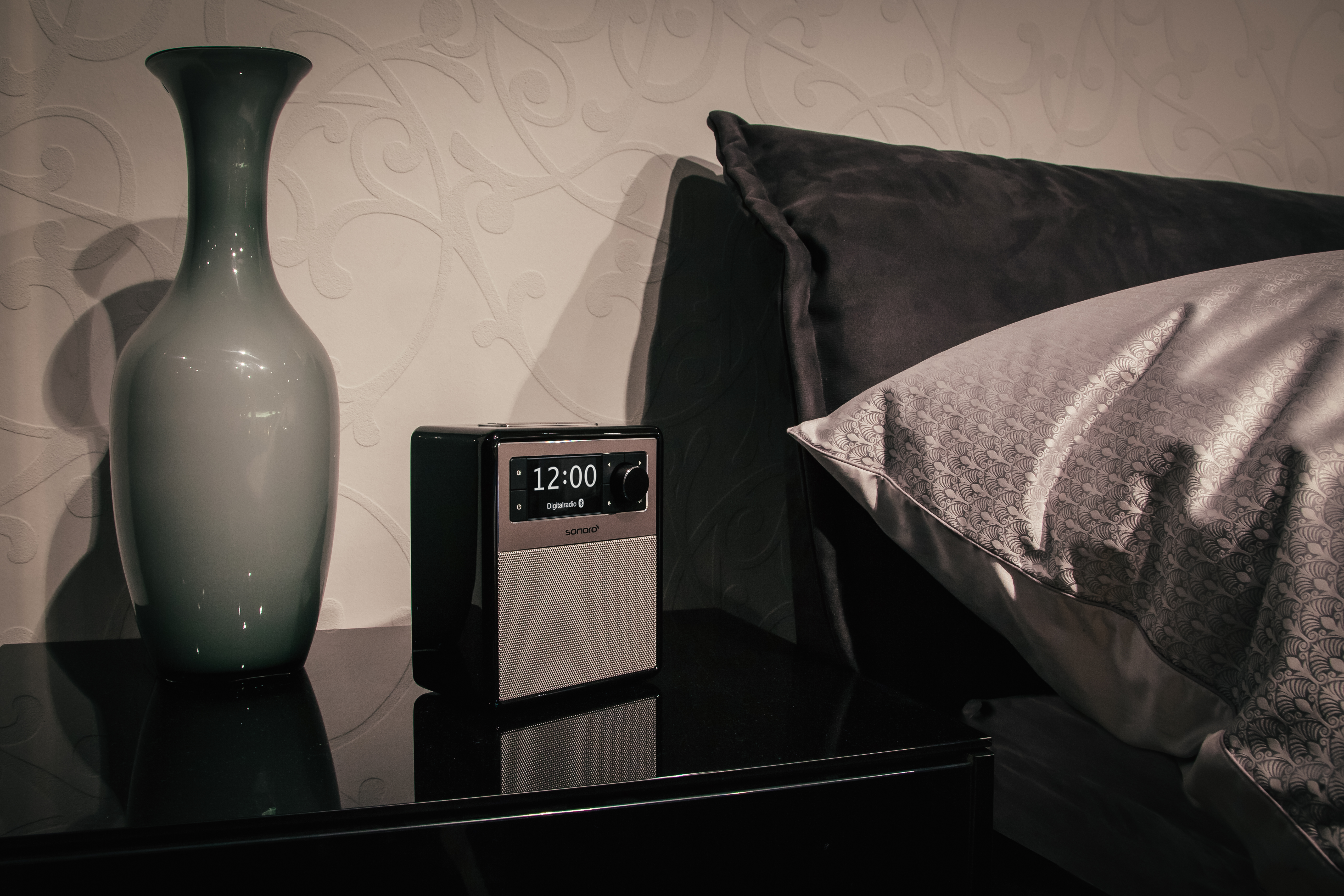 Radio auf einem Tisch neben einem Bett. Eine Vase ist links. Bettwäsche im Hintergrund.
