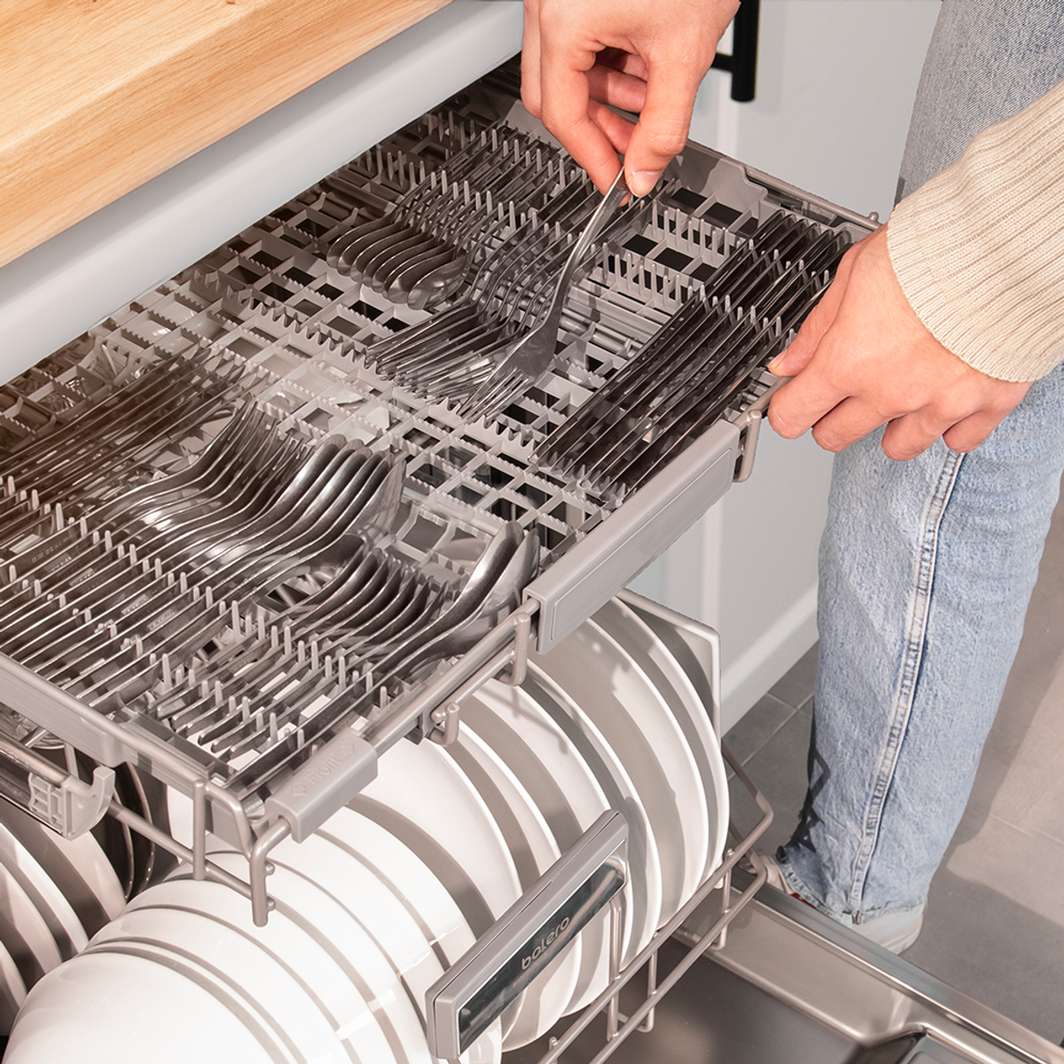Osoba ładuje sztućce do kosza zmywarki. Talerze widoczne na dolnej półce.