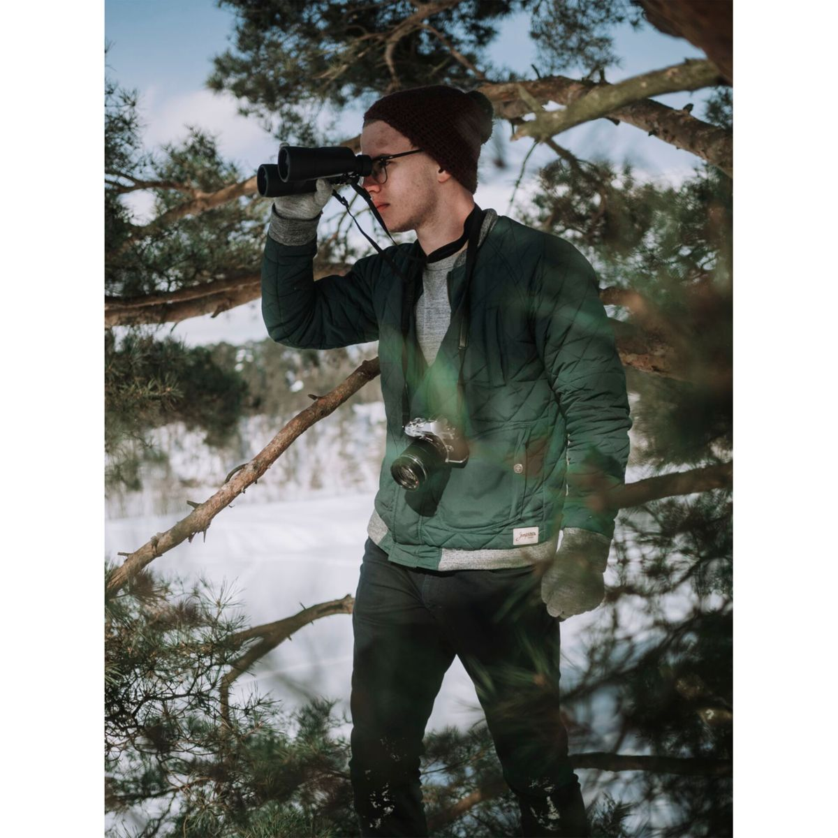 Mężczyzna w zielonej kurtce i okularach spogląda przez lornetkę w ośnieżonym lesie. Aparat na piersi.