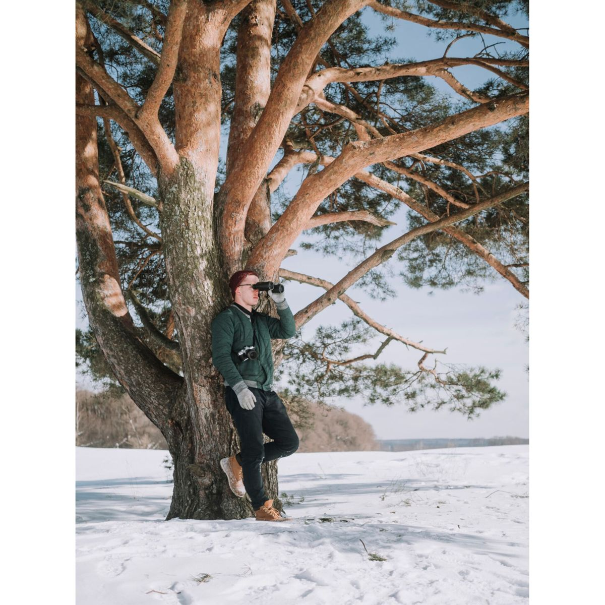 Mężczyzna z lornetką przy drzewie na zaśnieżonym polu, błękitne niebo.
