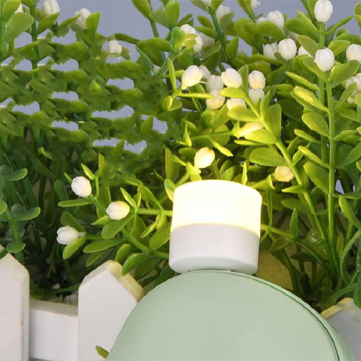 Ein kleines weiß-graues Licht vor einer grünen Pflanze mit weißen Blüten.