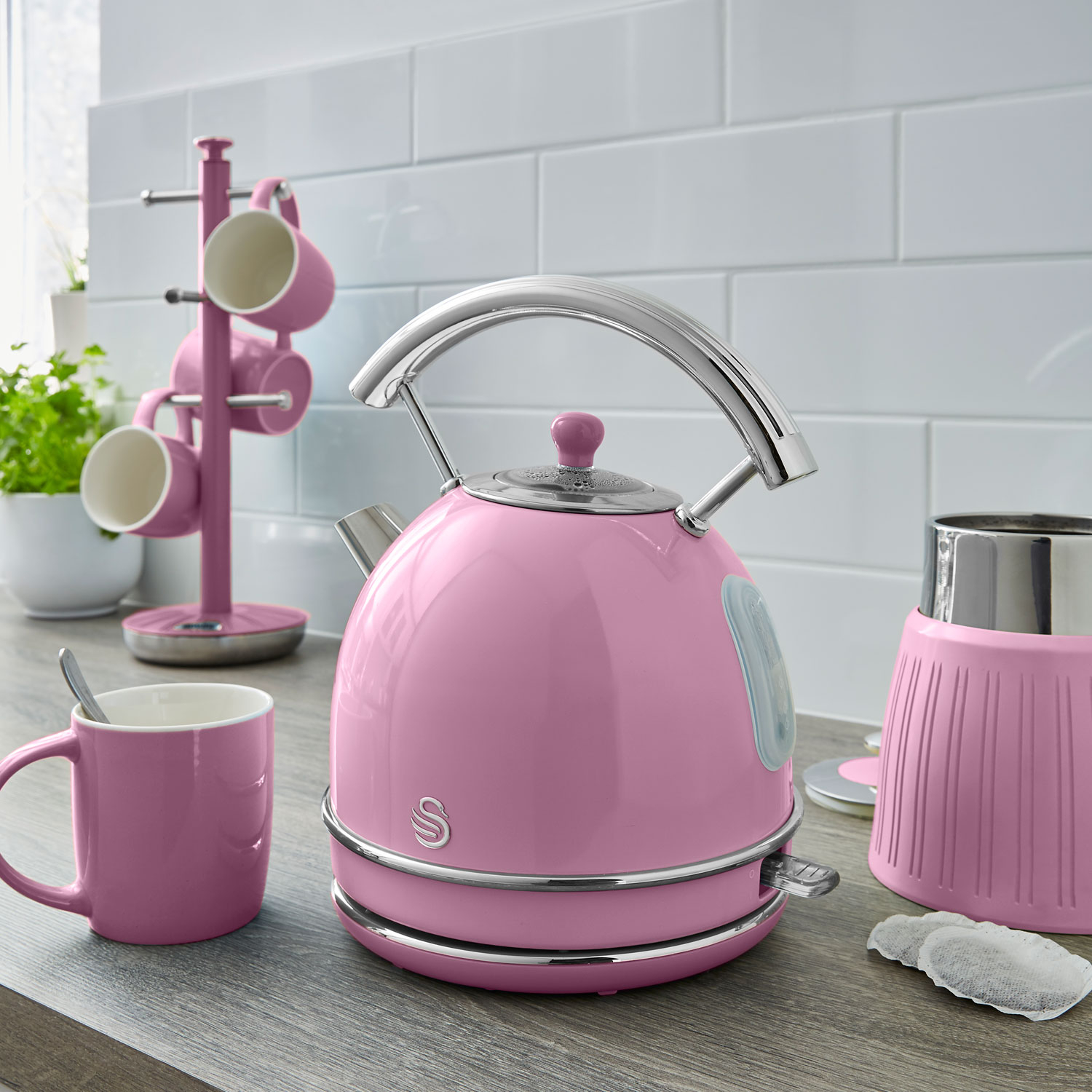Hervidor de agua rosa y accesorios sobre una encimera. Hay una taza, un soporte y bolsitas de té.