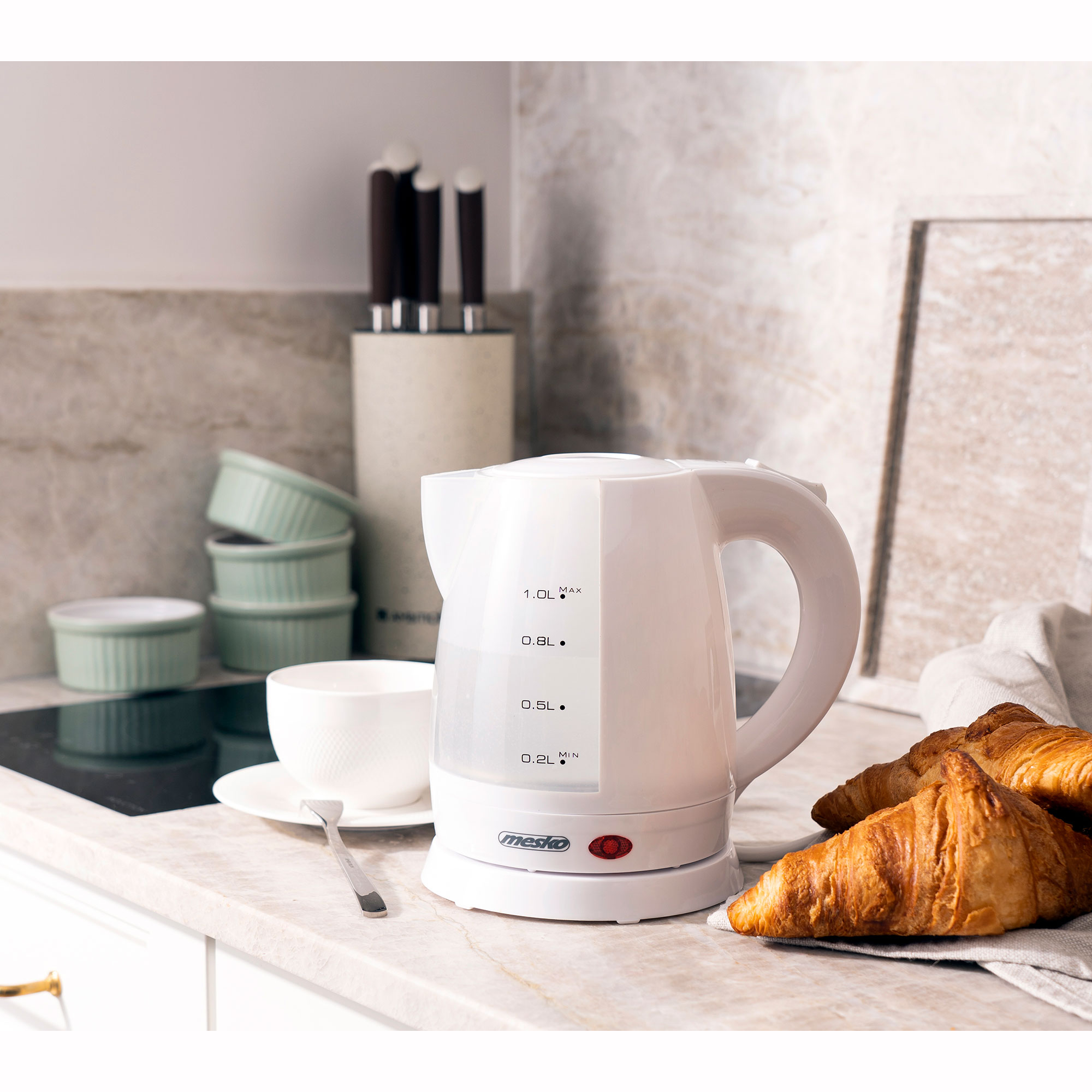 Hervidor eléctrico blanco en una encimera con taza, platillo y cruasanes en el fondo.