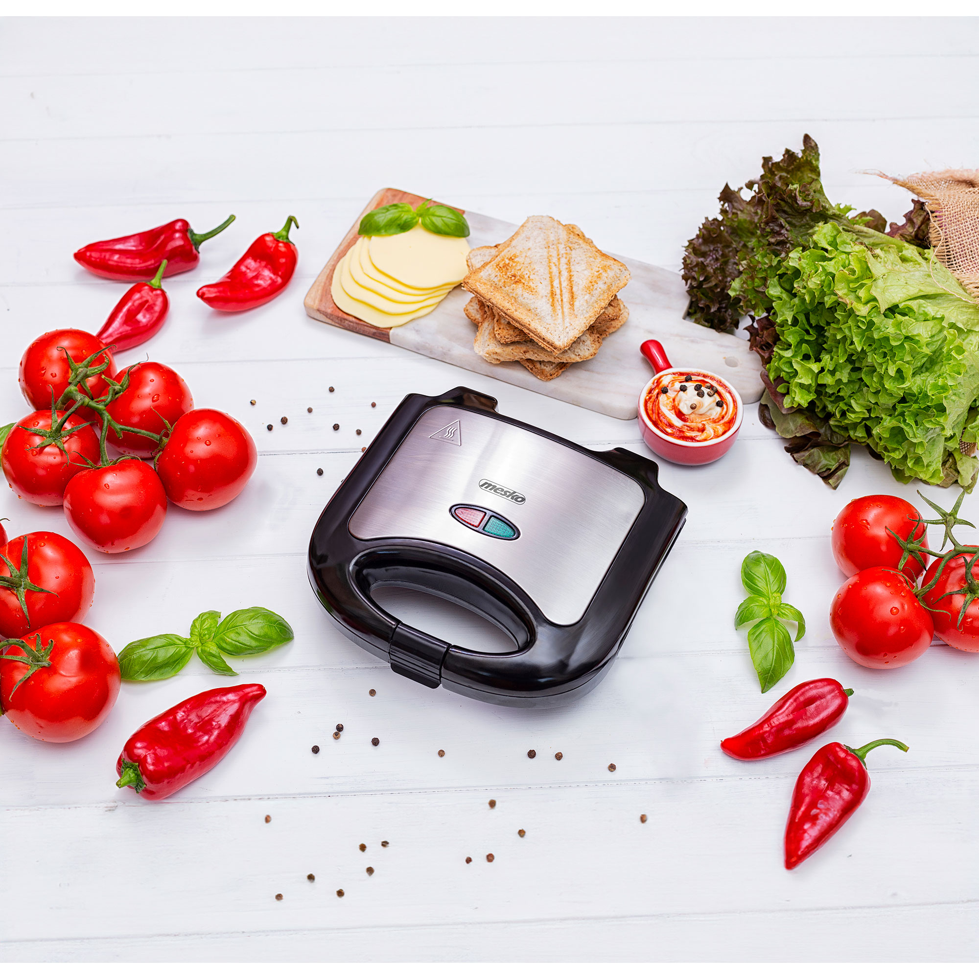Una sandwichera con ingredientes como tomates, queso y pan, sobre una superficie blanca.
