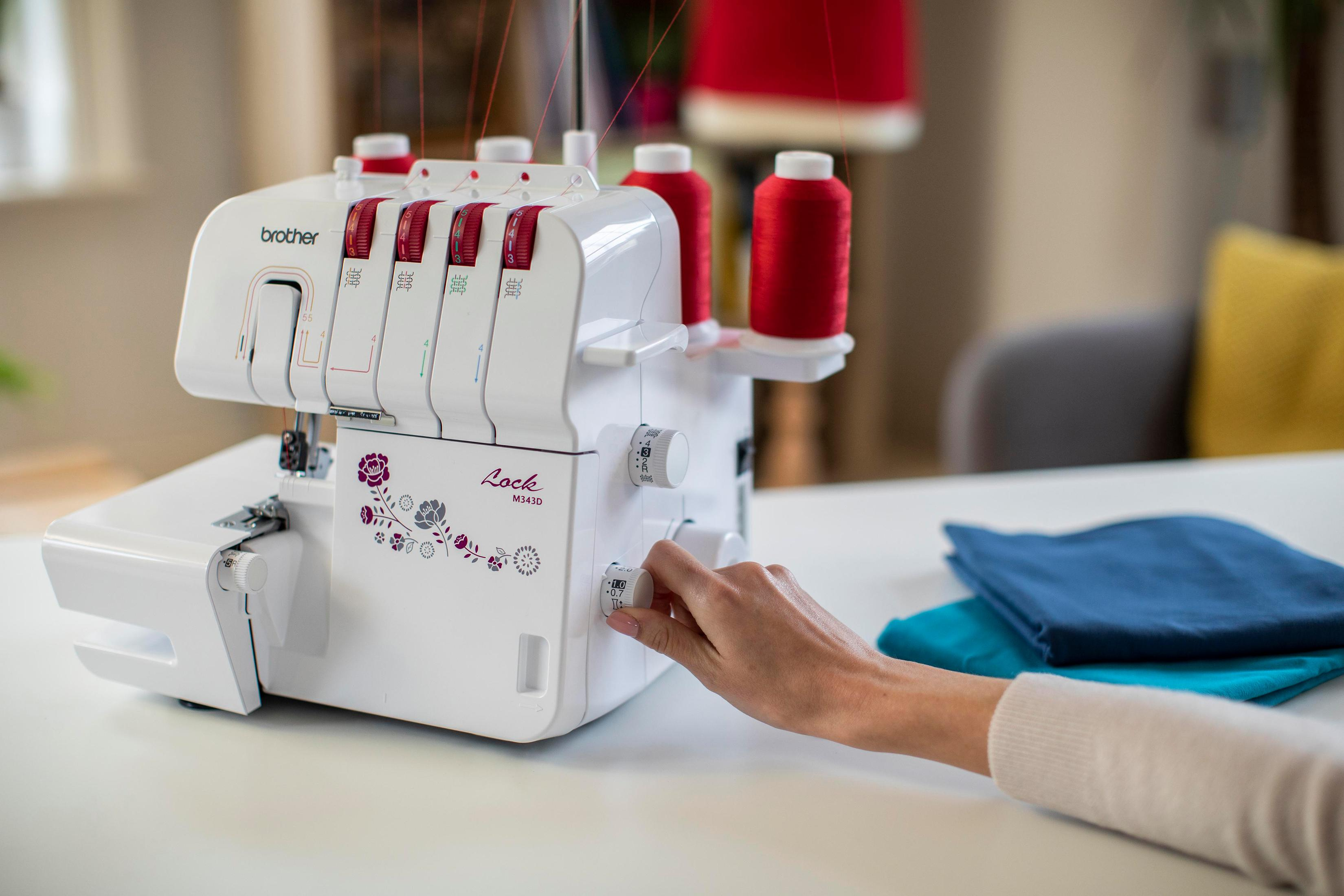 Biała maszyna do szycia z czerwoną nicią. Ręka reguluje pokrętło. Niebieskie i turkusowe tkaniny na białym stole.