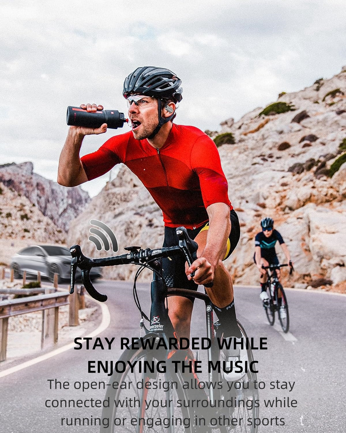 Ciclista bebiendo de una botella, usando auriculares. Otro ciclista al fondo. Carretera y montañas.