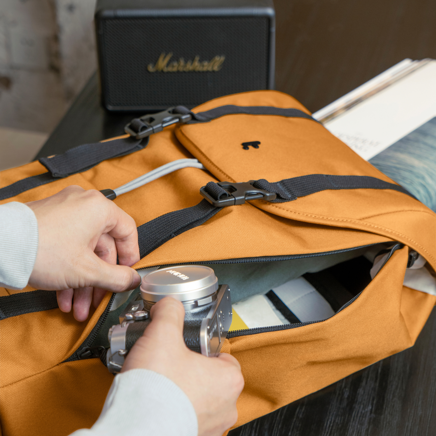 Handen openen een bruine tas, onthullend een camera erin, met een Marshall speaker achter.