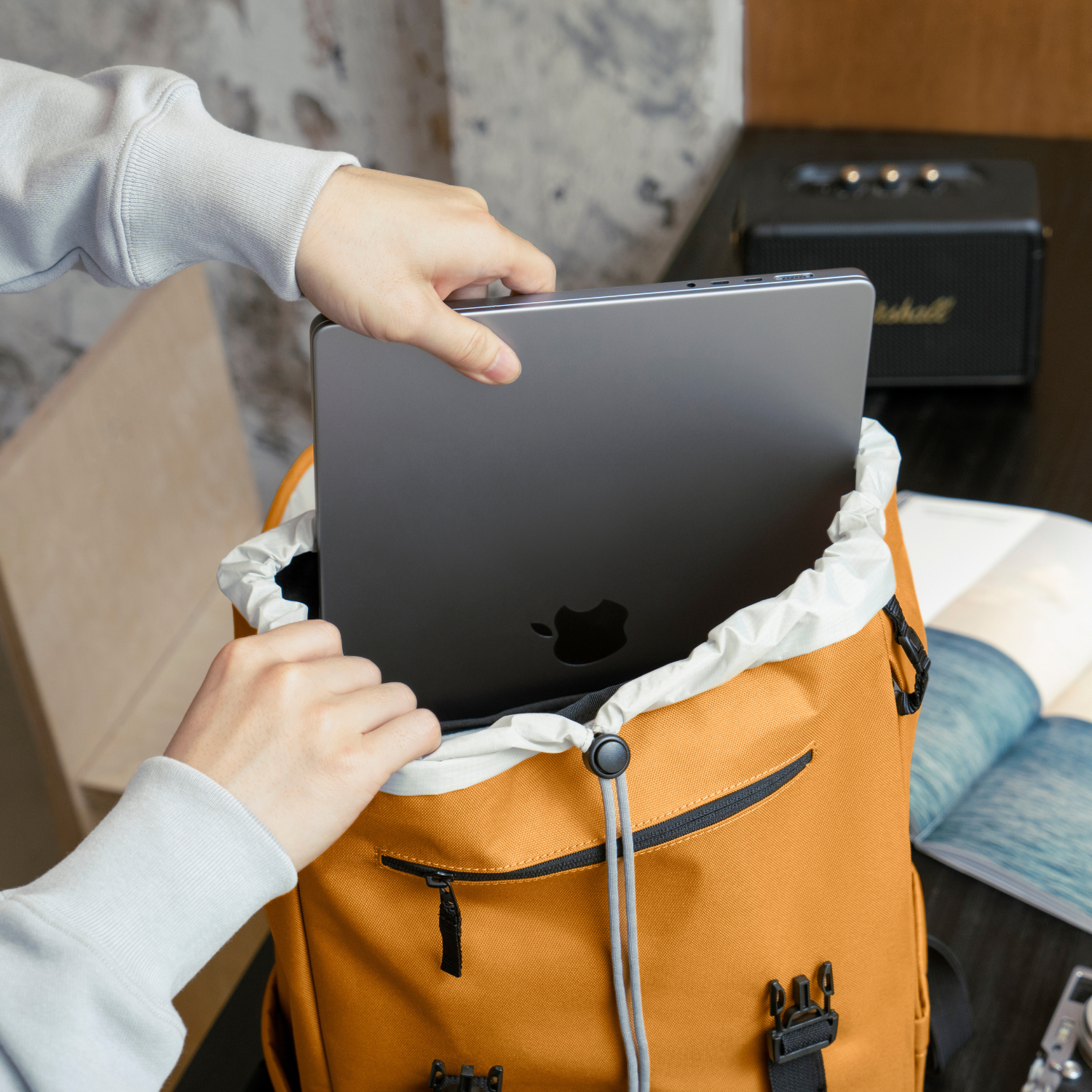 Een persoon die een laptop in een oranje rugzak stopt. Een zwarte speaker op de achtergrond.