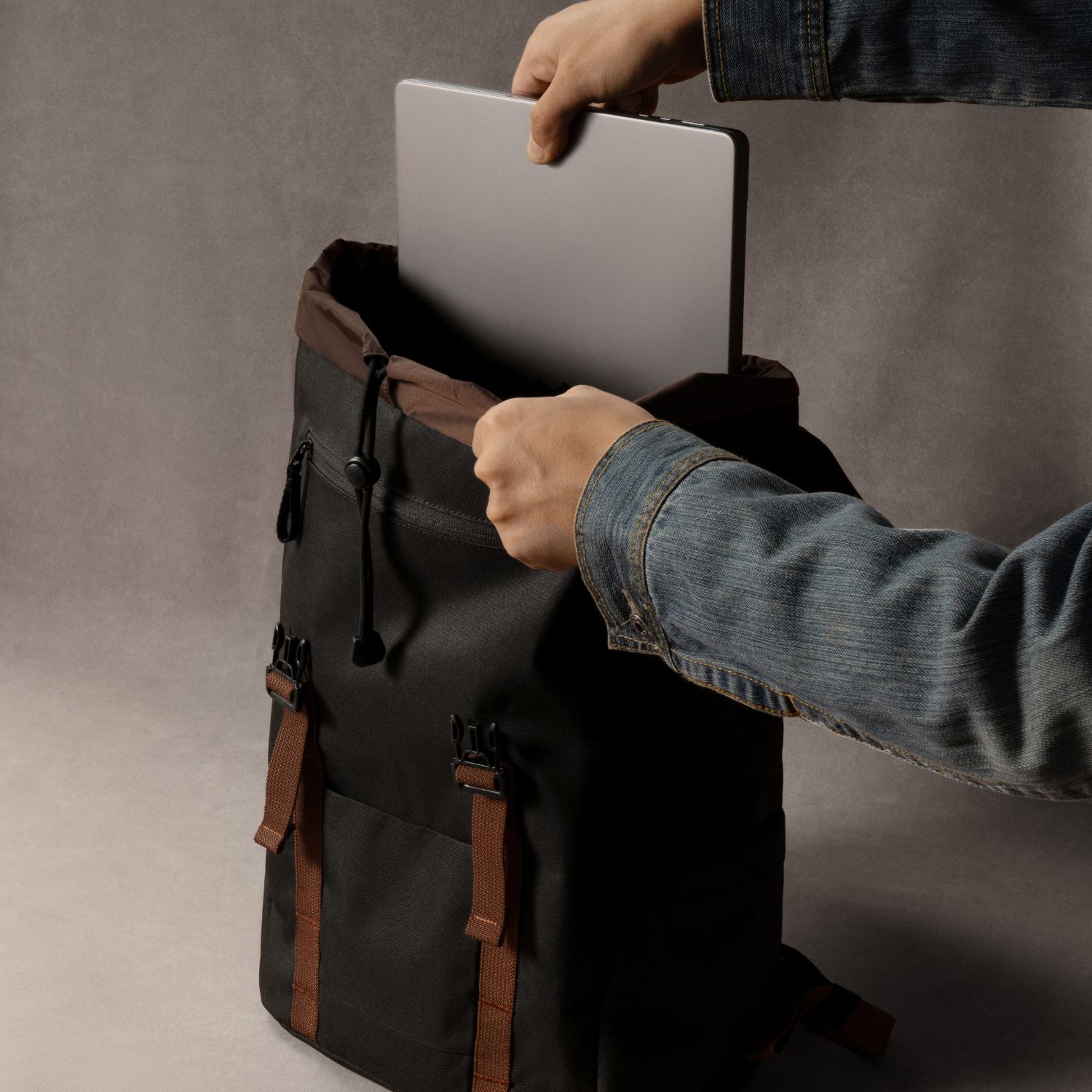 Ręce wkładają laptopa do czarnego plecaka. Osoba ma na sobie dżinsową kurtkę.