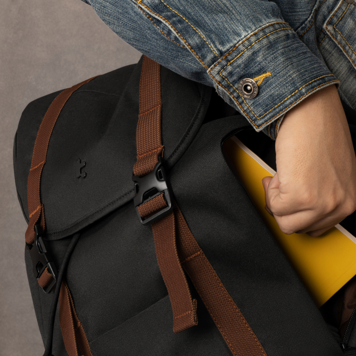 Ręka osoby sięgająca do czarnego plecaka. Wyciągany jest żółty notatnik.