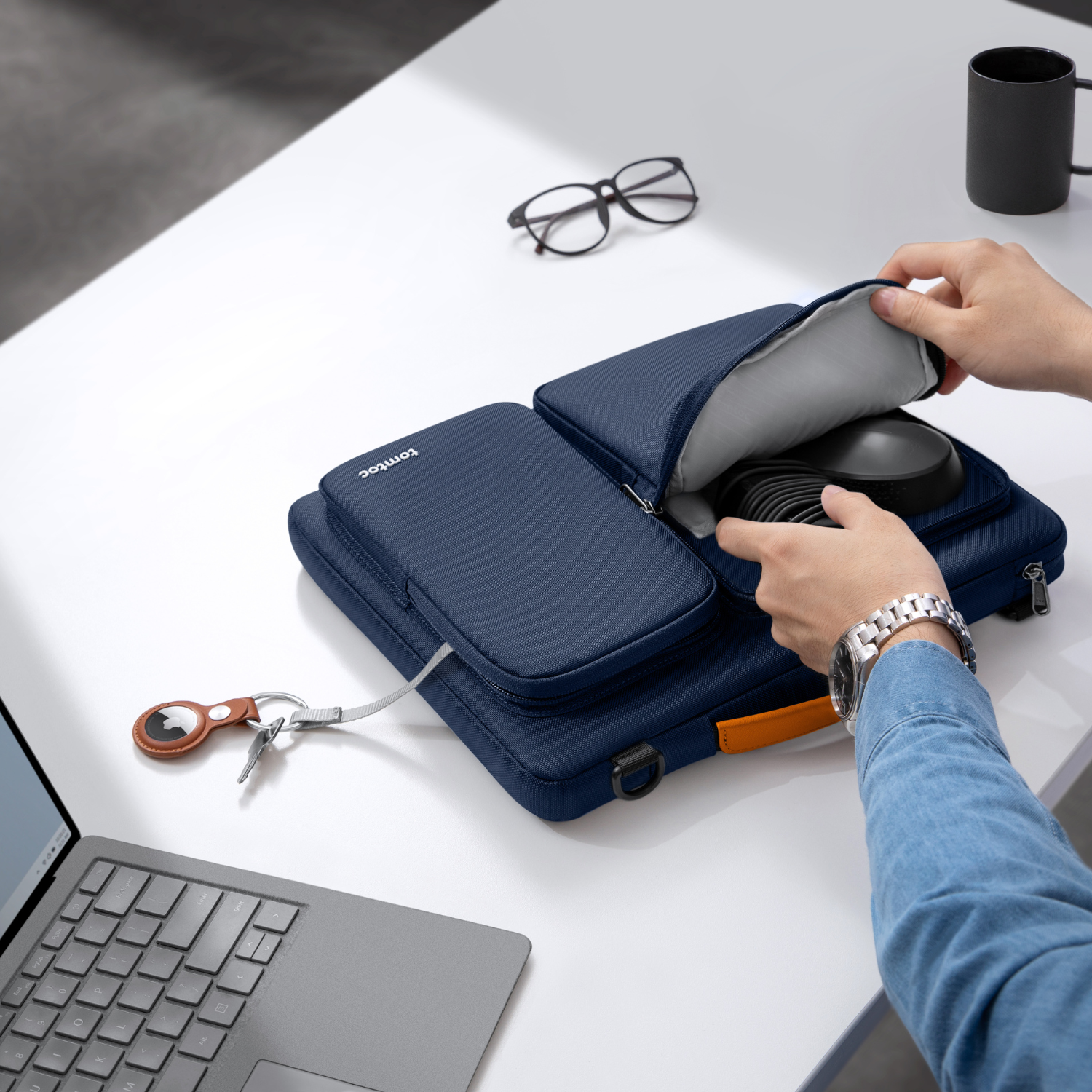 Mani che aprono una borsa blu per laptop tomtoc su una scrivania bianca, rivelando un laptop e accessori.