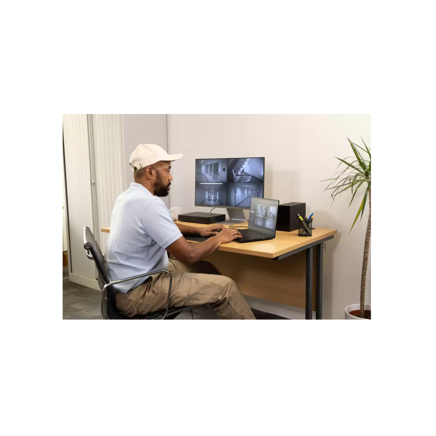Mężczyzna przy biurku z monitorem pokazującym nagrania z monitoringu i laptopem. Roślina i szafa w tle.