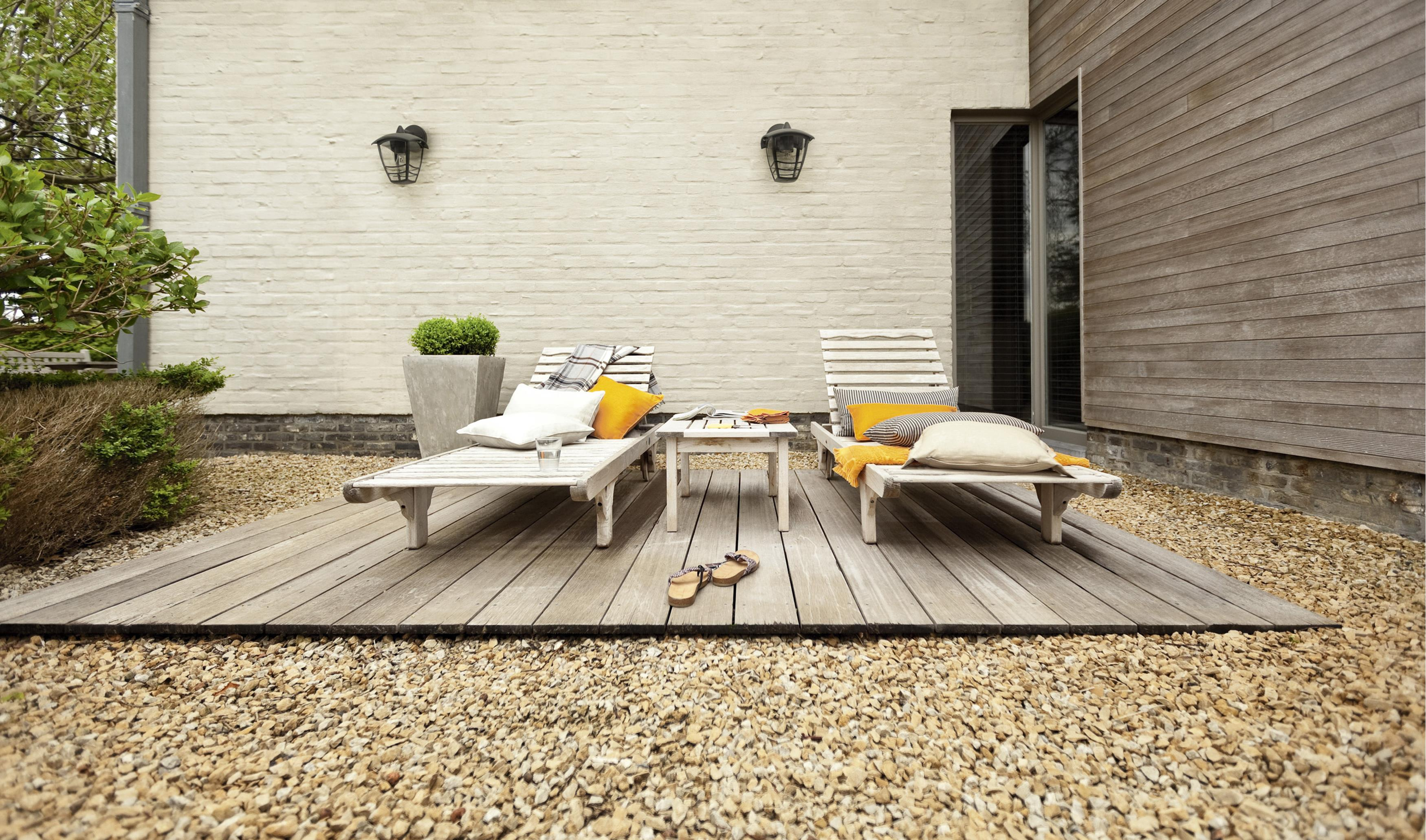 Eine Holzterrasse mit zwei Liegestühlen und einem kleinen Tisch. Der Hintergrund ist eine Backsteinmauer.