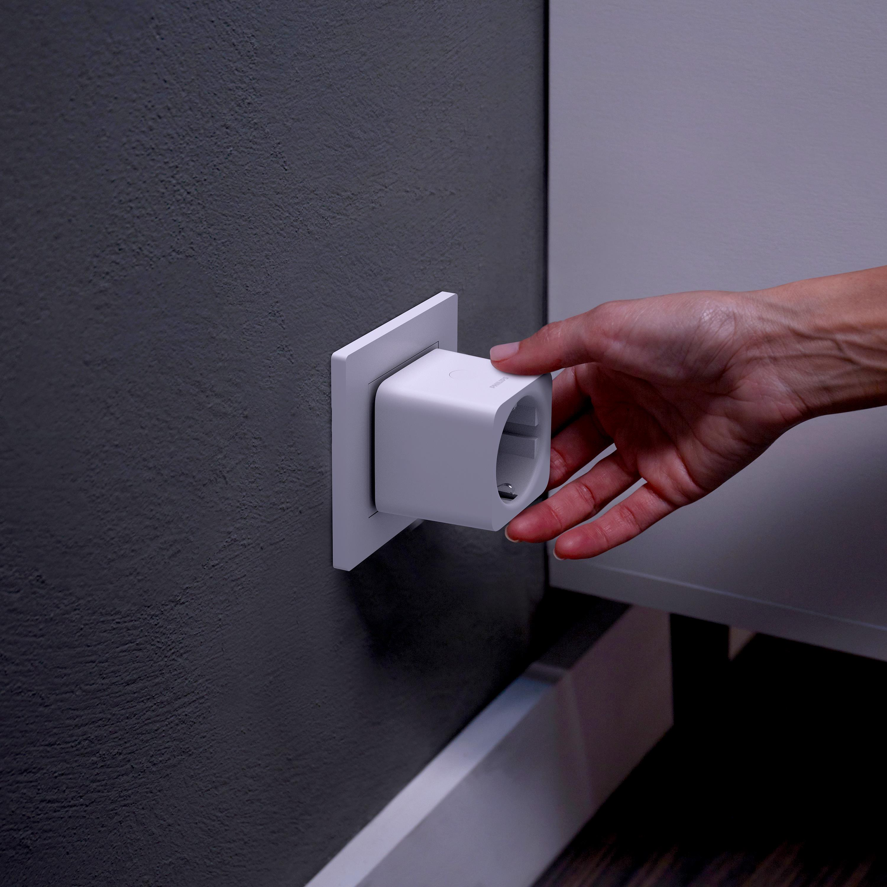 A hand plugs a white smart plug into a grey wall outlet.