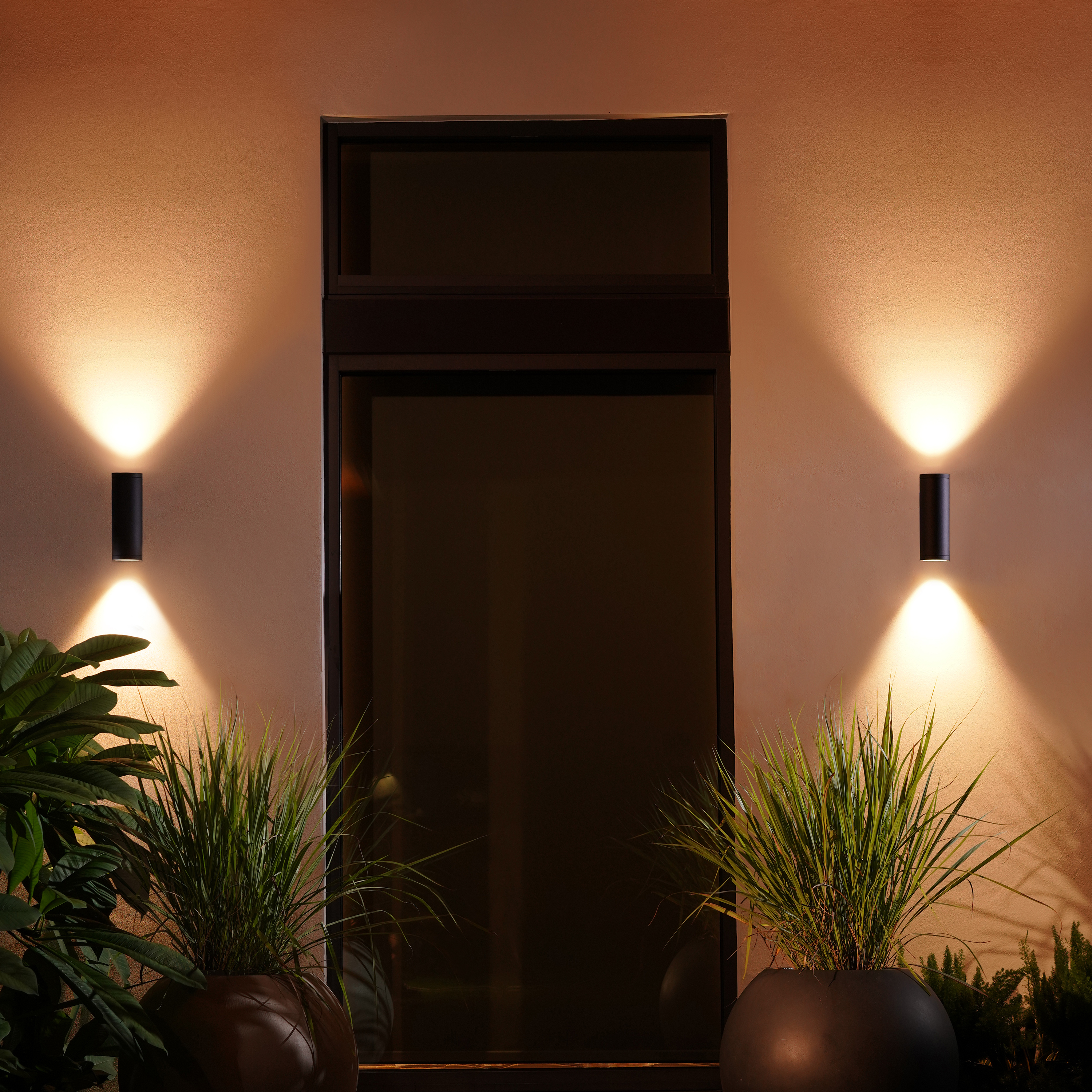 Mur extérieur avec deux lumières cylindriques noires, éclairant vers le bas. Plantes en pots en dessous.