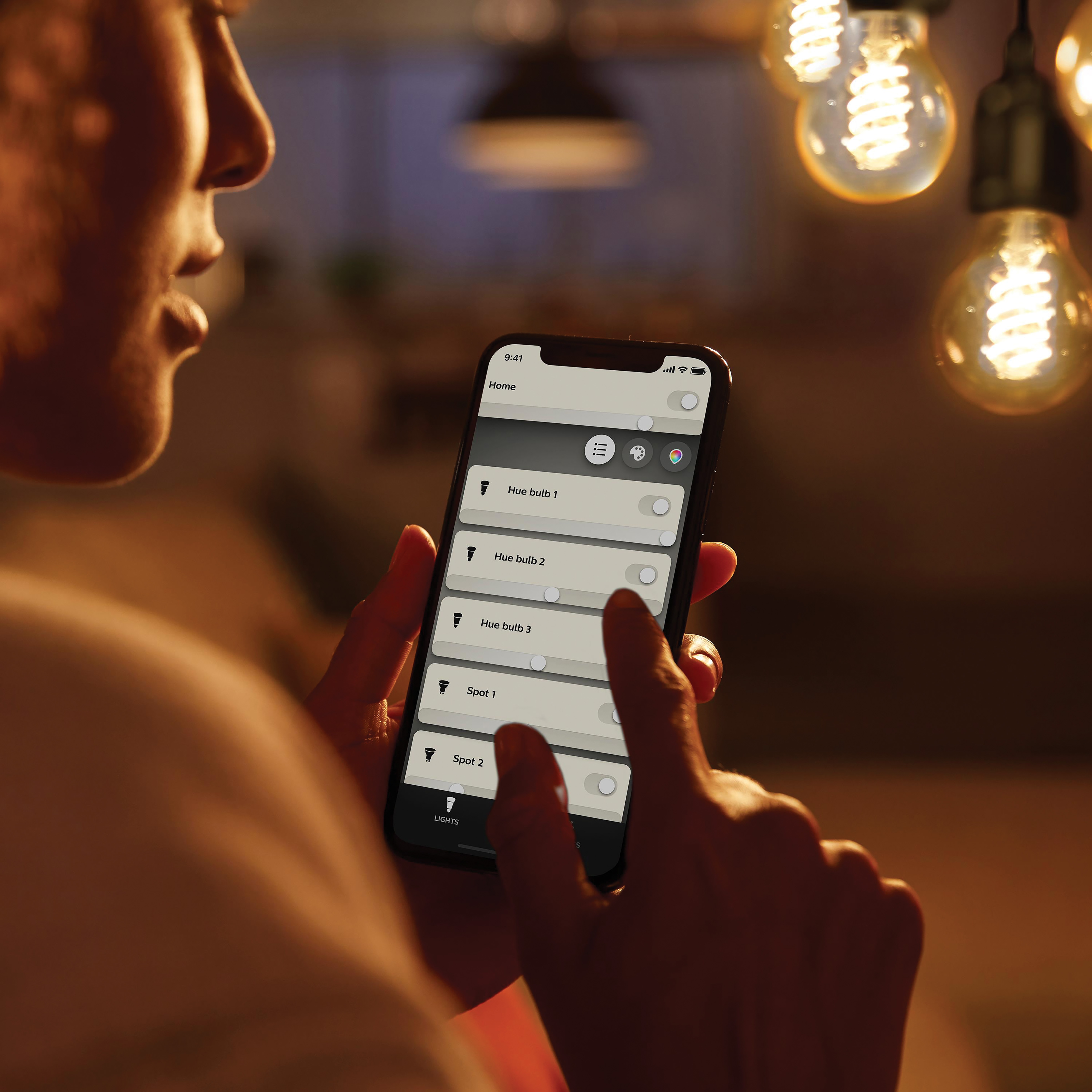 Eine Person steuert Smart-Glühbirnen mit einem Smartphone. Mehrere Glühbirnen hängen darüber.