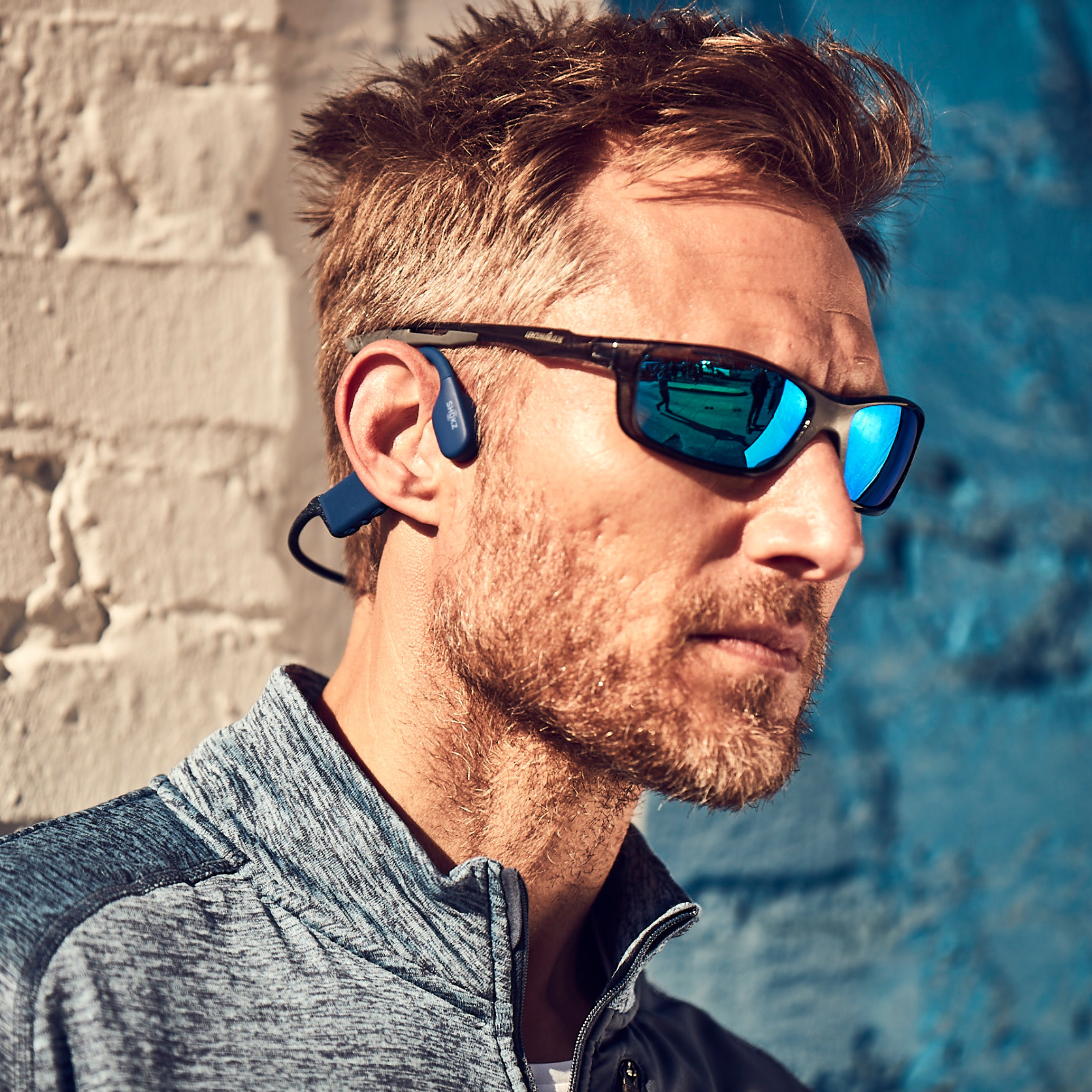 Mężczyzna w okularach przeciwsłonecznych i niebieskich słuchawkach opiera się o ceglany mur. Ma krótką brodę.